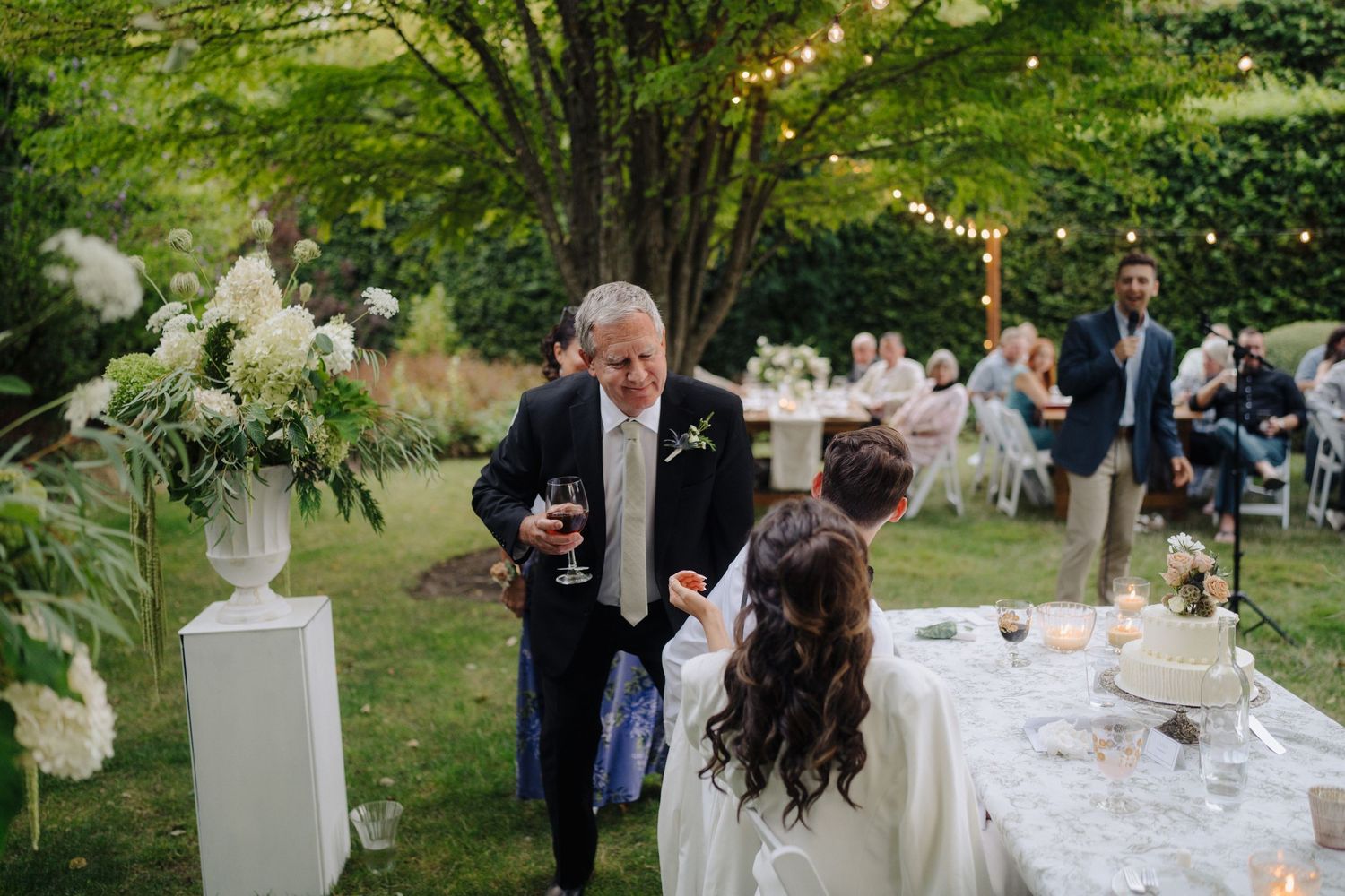 Wedding reception dining tables decorated with white linens and floral arrangements under trees.