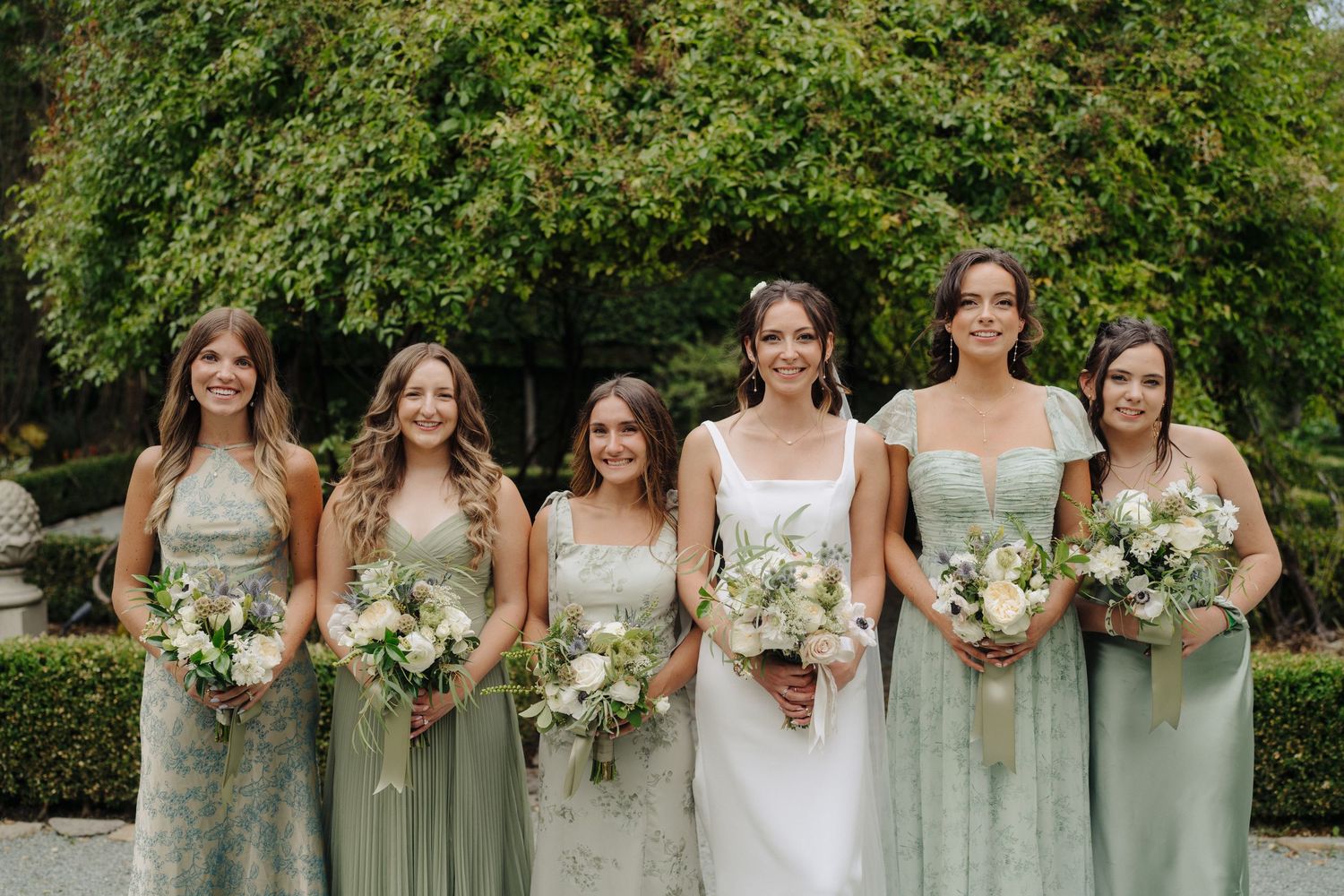 Bridesmaids in sage green dresses holding white bouquets pose against green foliage.