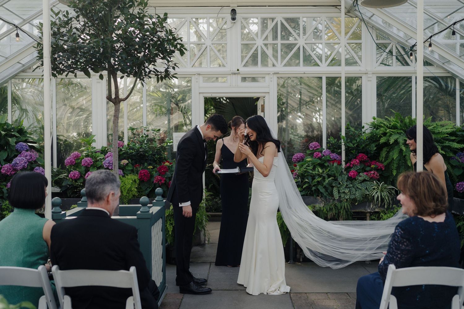 A wedding ceremony takes place in a beautiful greenhouse with white decor and florals.