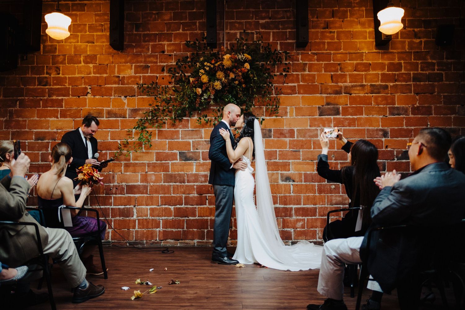 Couple shares their first kiss as newlyweds against a rustic brick wall backdrop.