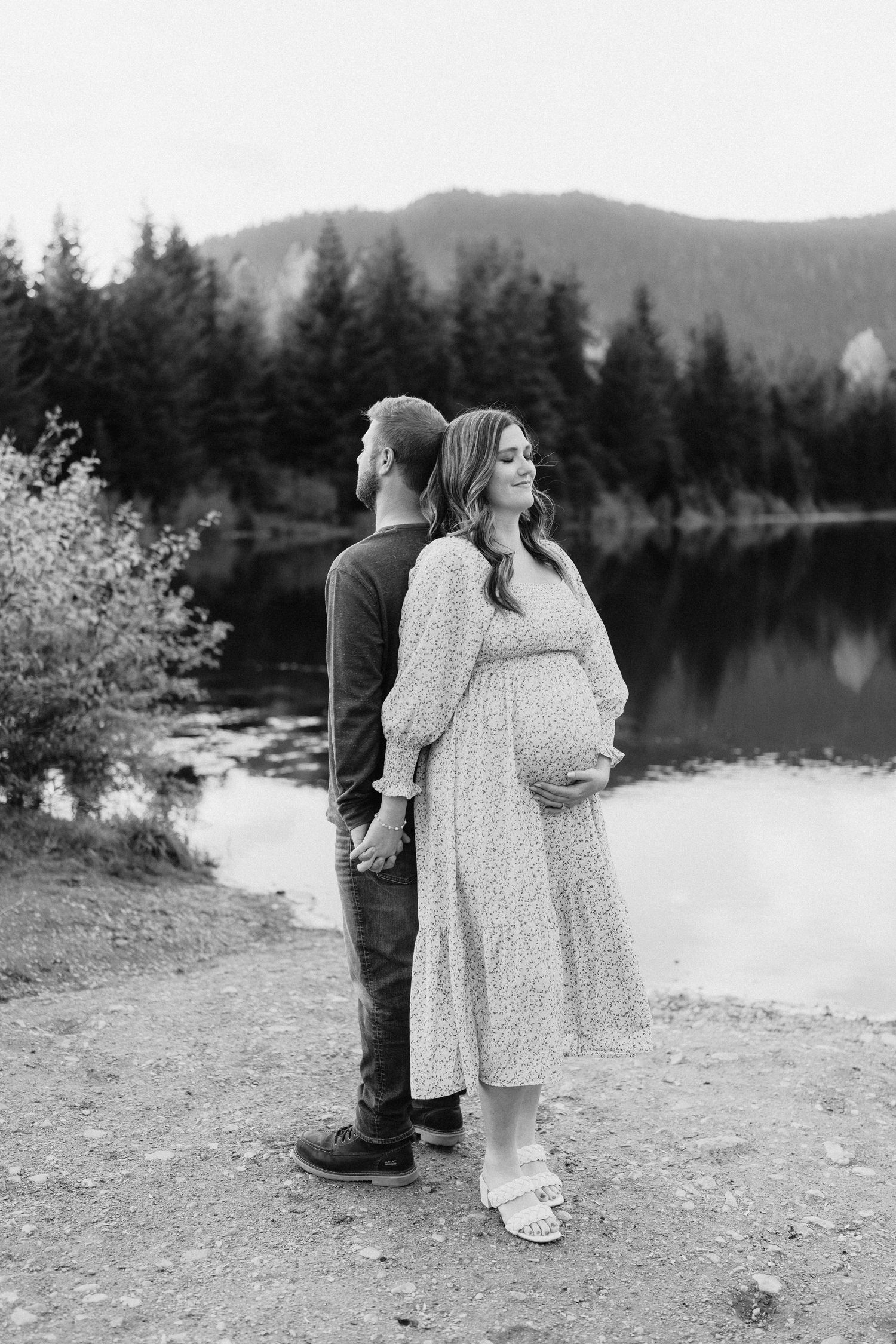 A couple stands together by a serene mountain lake surrounded by evergreen forest in black and white.