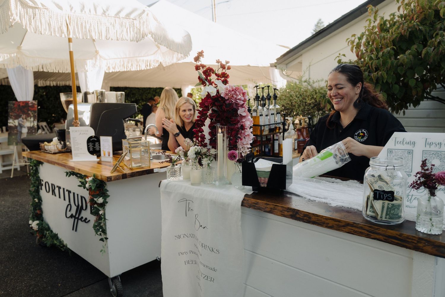 White reception bar setup with floral arrangements and glassware under tent lighting.