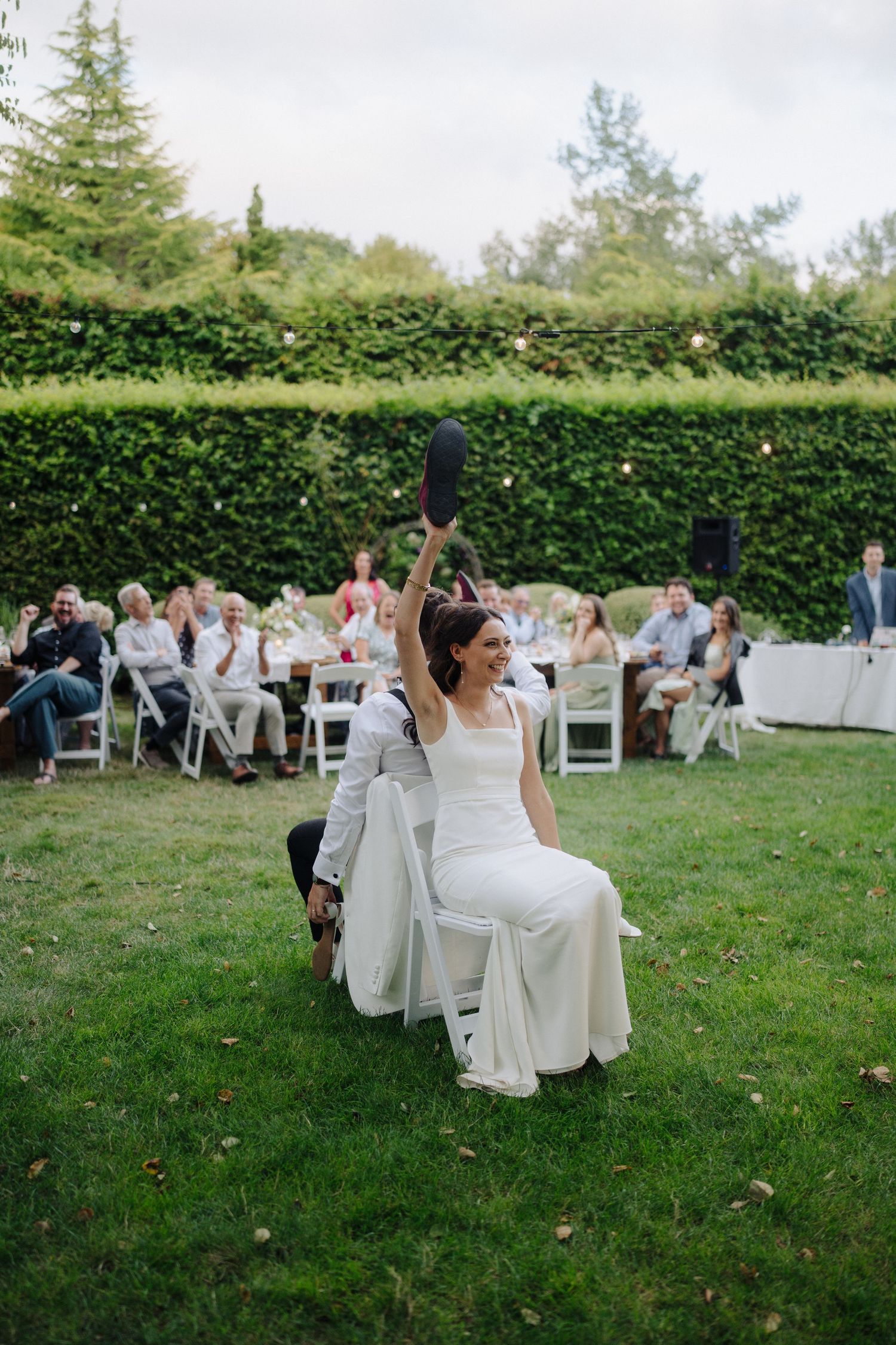 Wedding guests seated on white chairs participate in an outdoor game on a grassy lawn.