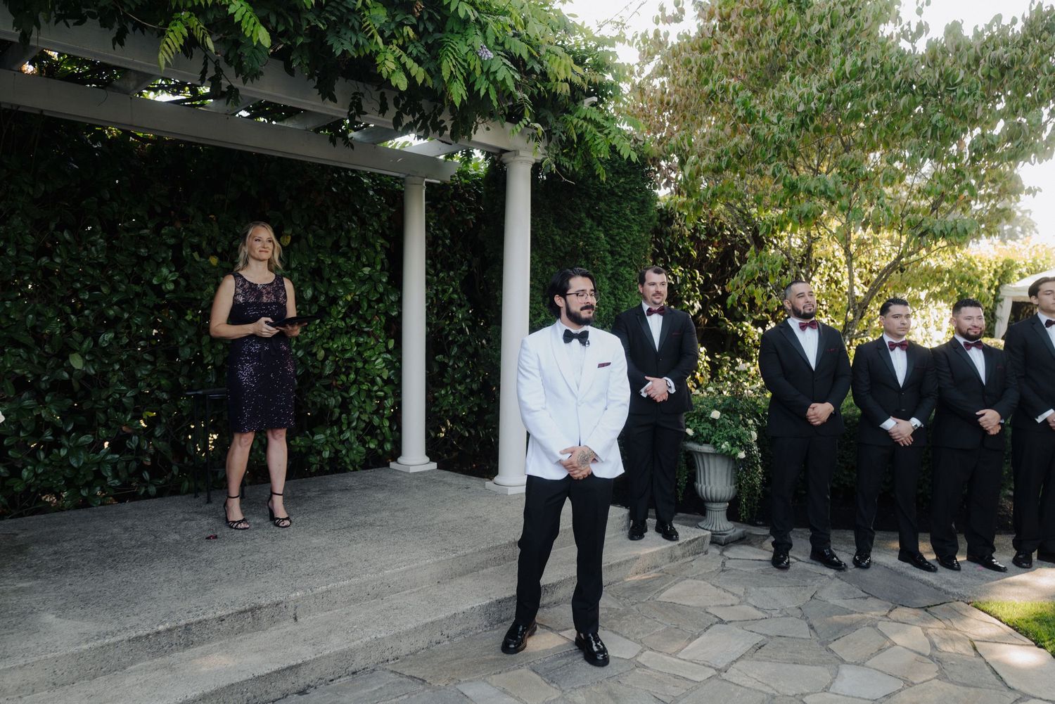 Wedding guests in formal attire walking down an outdoor aisle lined with white chairs.