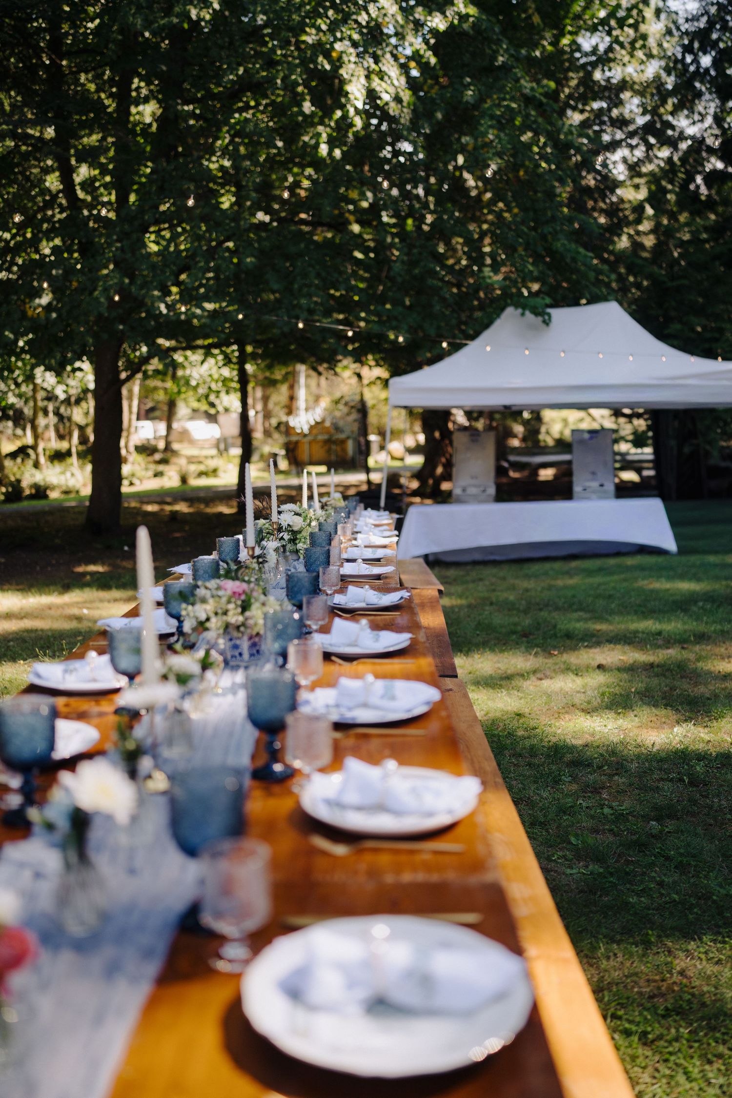 Long wooden table with white place settings arranged outdoors under trees for wedding reception.
