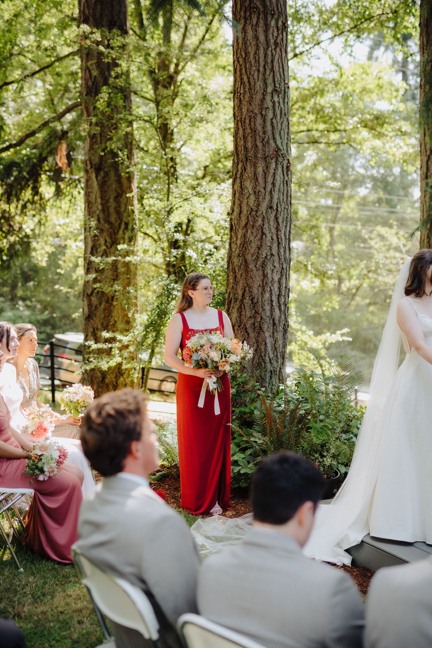 Wedding guests watch ceremony in outdoor woodland setting with sunlight filtering through tall trees.