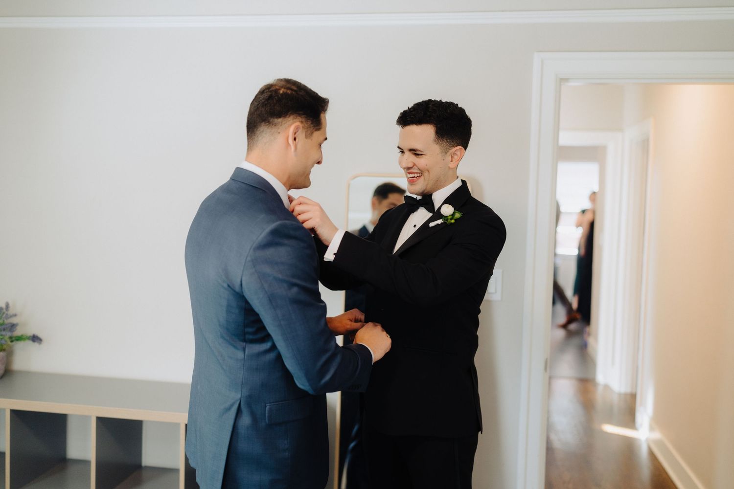 Two people in formal suits share a heartfelt moment while adjusting each other's bow ties before a wedding.