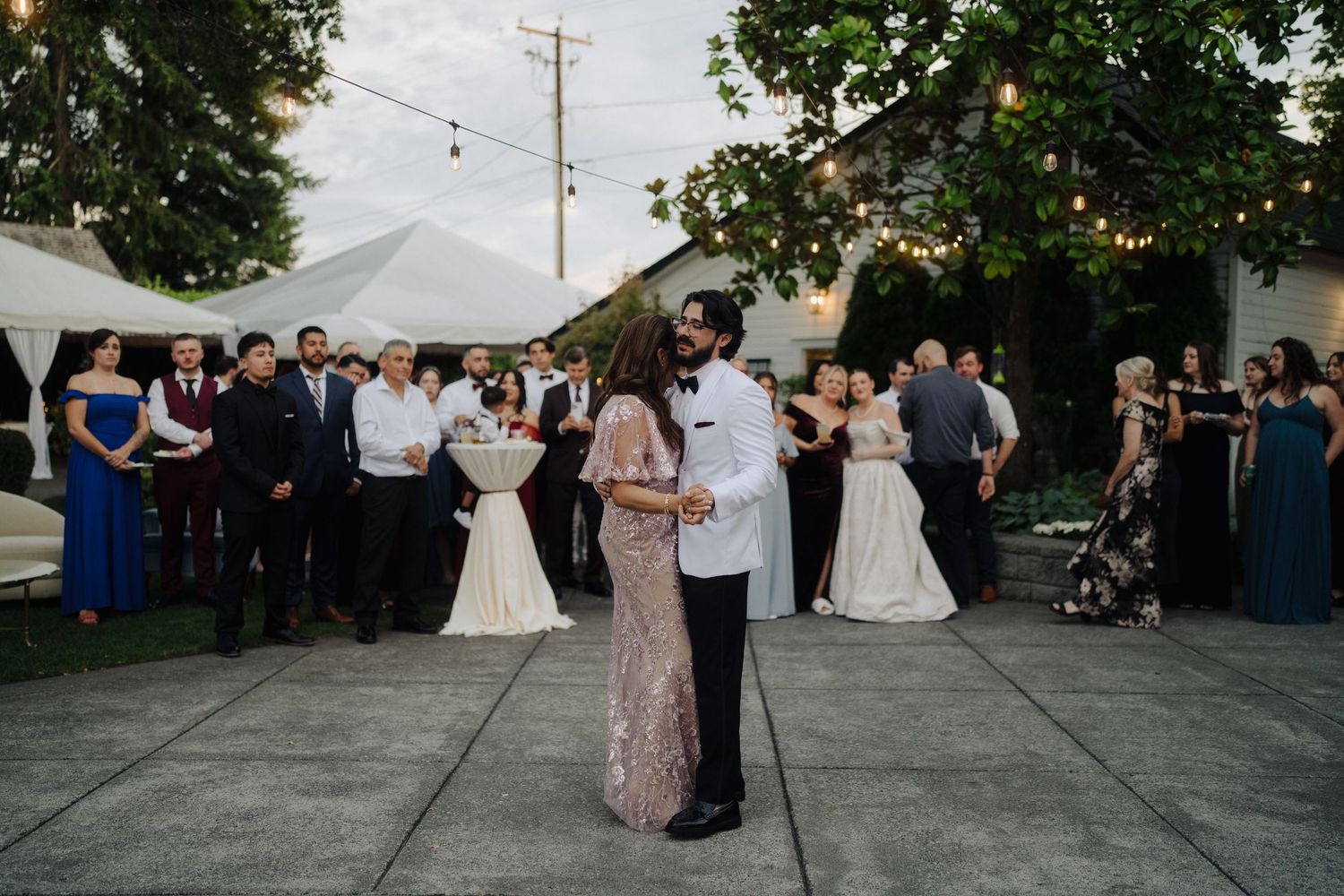 Outdoor wedding reception with string lights and dancing guests at dusk.