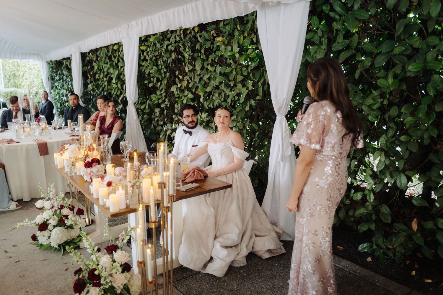 An elegant wedding reception setup with candlelit tables and greenery wall backdrop.