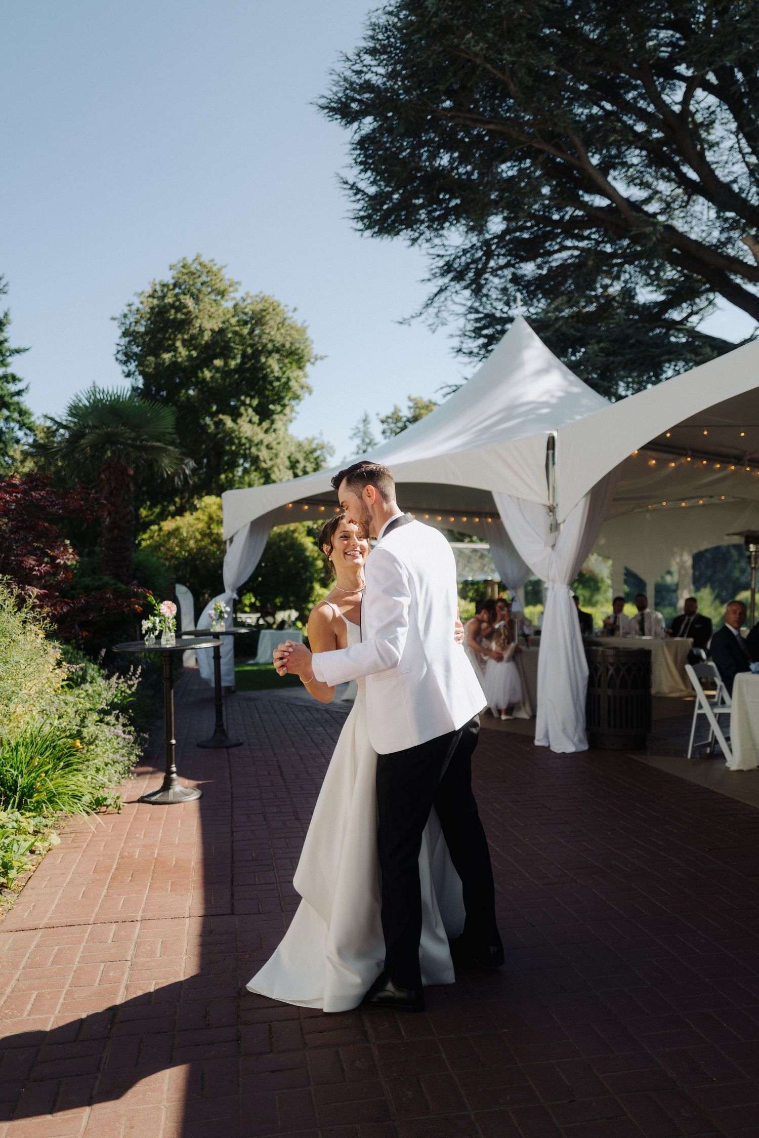 Outdoor wedding dance under white tent with brick patio and trees.