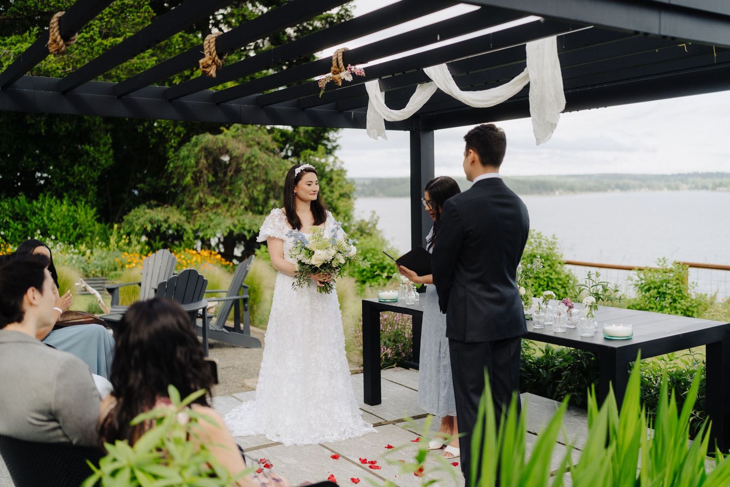 An intimate outdoor wedding ceremony takes place under a pergola with lush greenery and waterfront views.