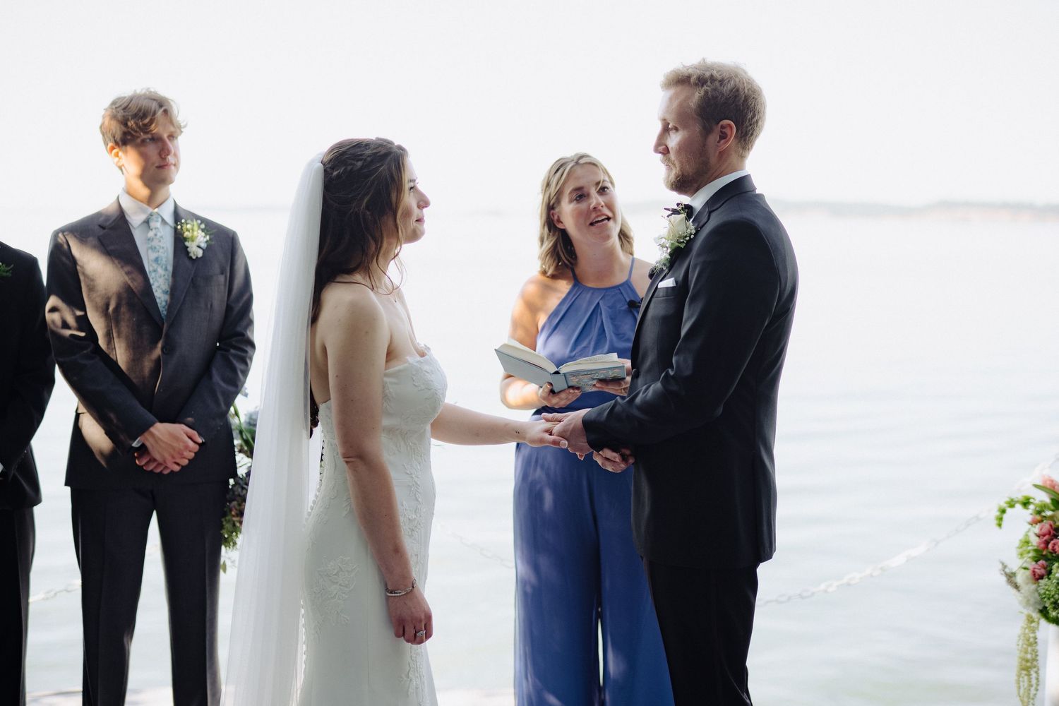 Wedding ceremony takes place against a scenic waterfront backdrop with guests in formal attire.