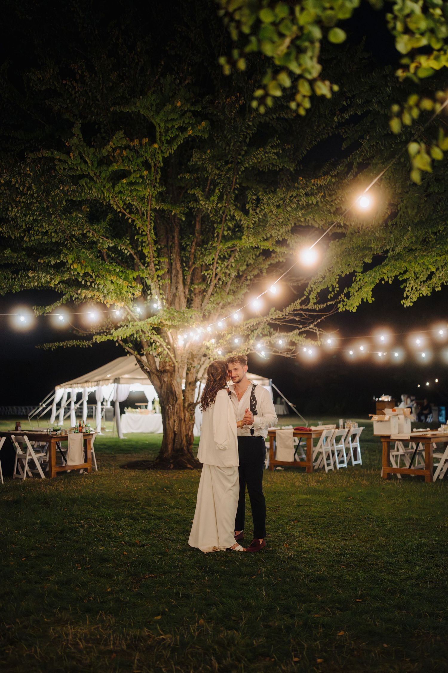 String lights illuminate an outdoor evening wedding reception under large trees.