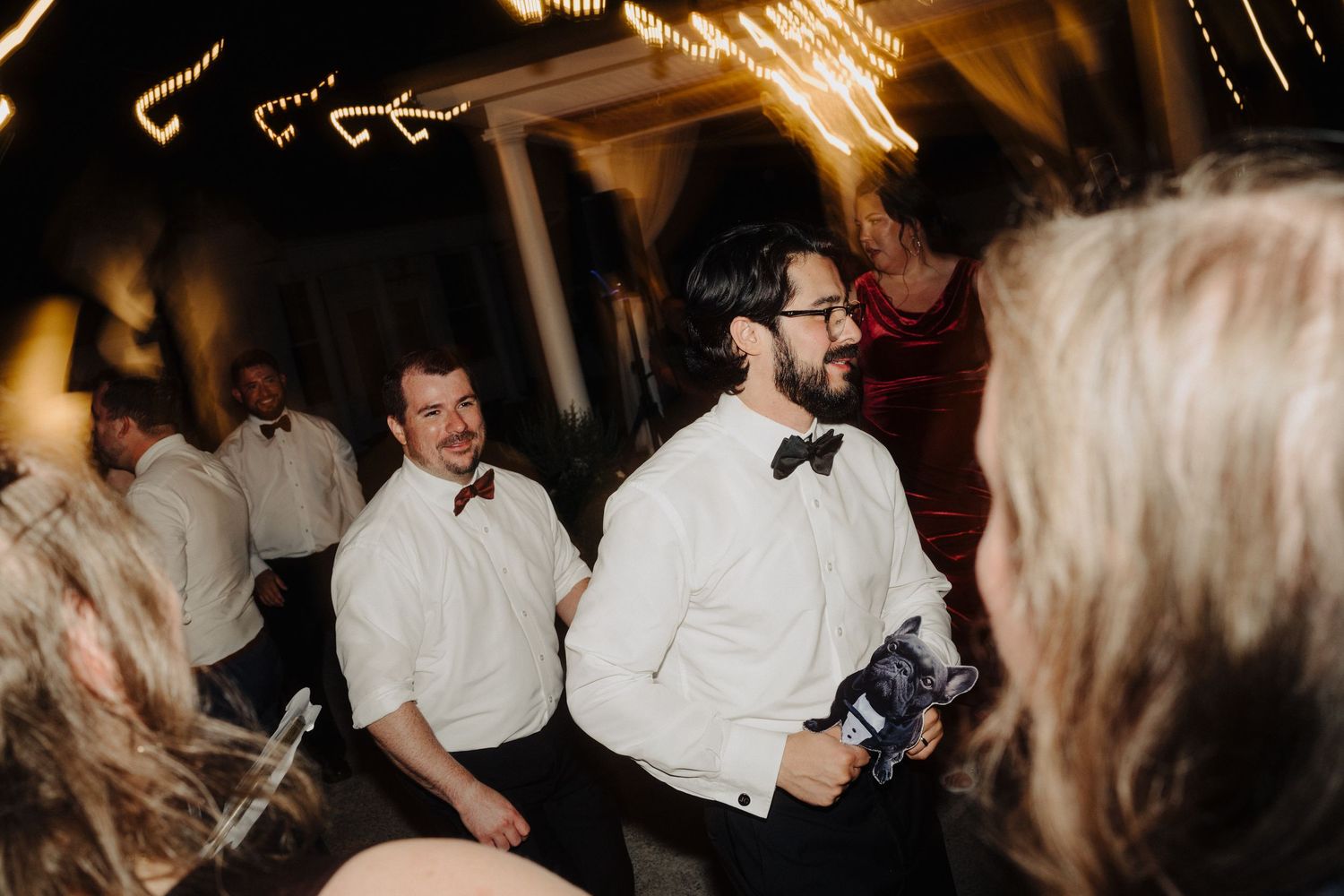 Wedding guests dancing and celebrating on a dimly lit dance floor with string lights overhead.