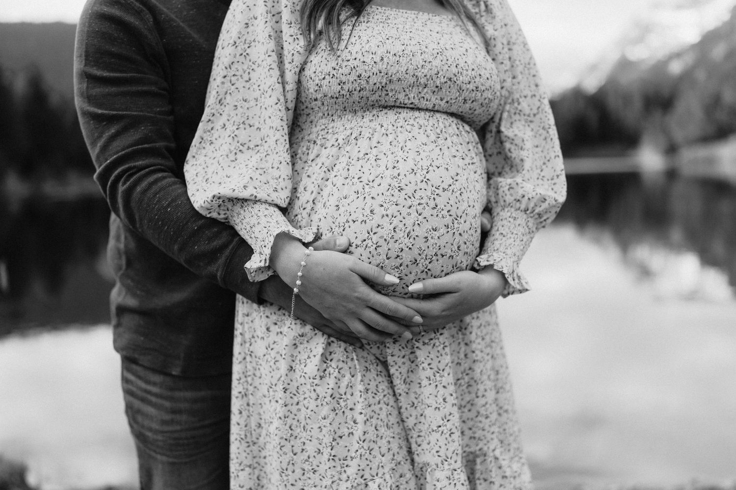A couple in a floral dress and dark shirt embrace while standing outdoors during a maternity photoshoot.