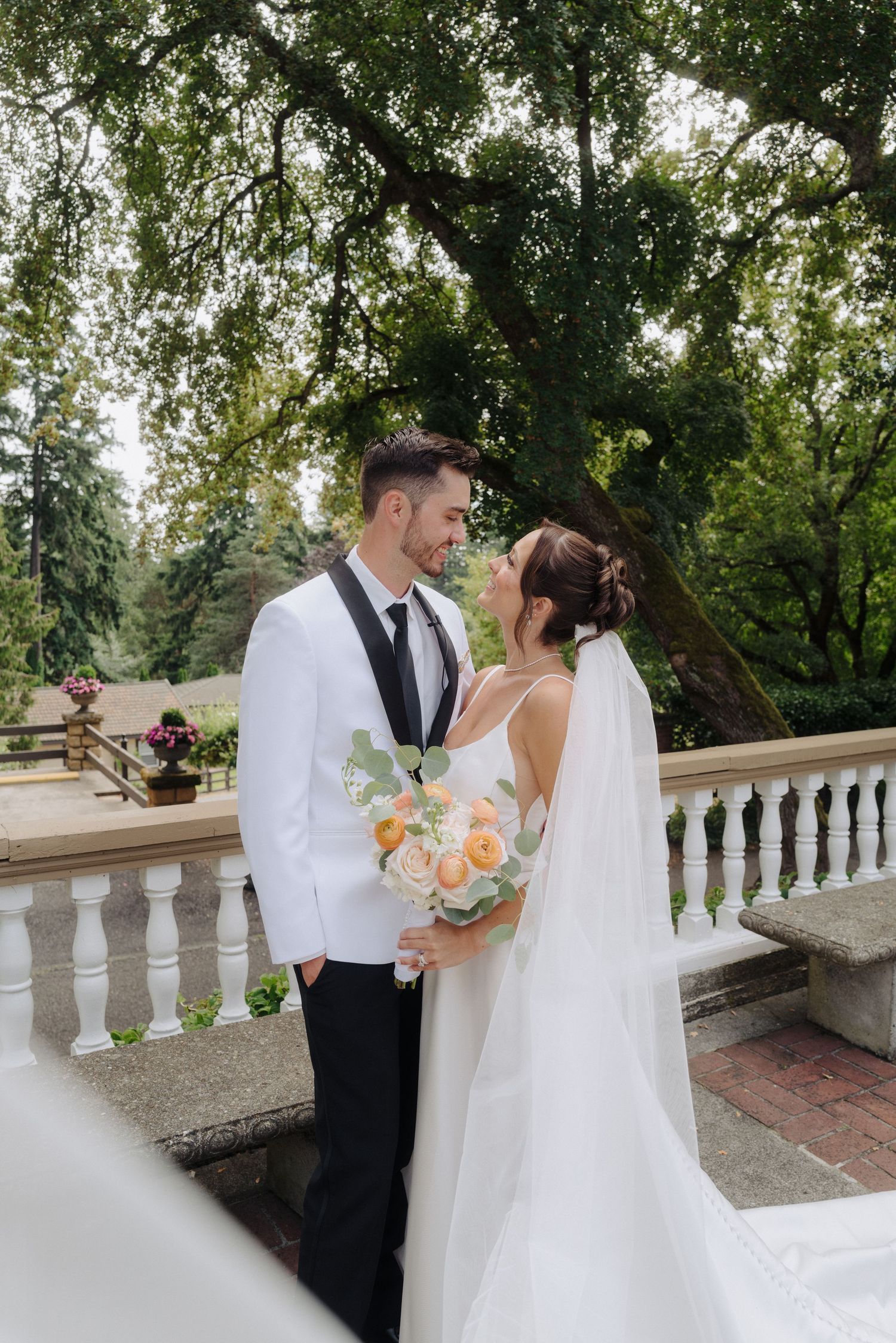 A romantic wedding couple shares a moment on a balcony with white railings surrounded by lush greenery.