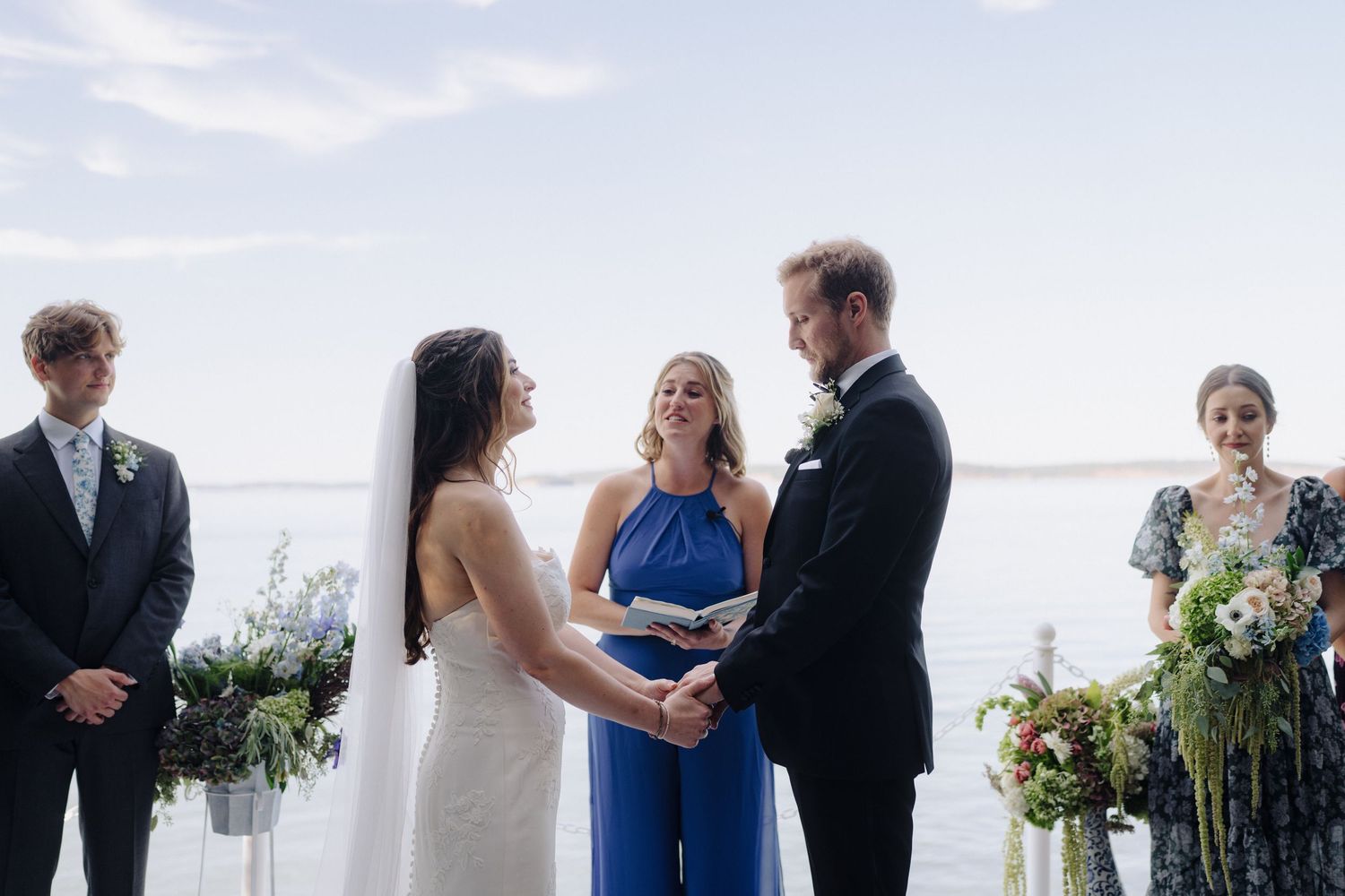 Intimate outdoor wedding ceremony with officiant in blue dress conducting service by waterfront.