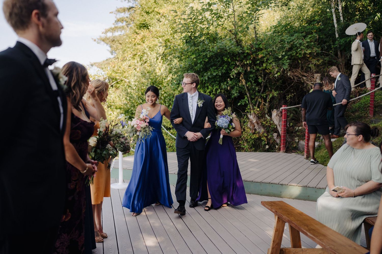 A wedding ceremony takes place on a wooden deck overlooking a scenic waterfront view at sunset.