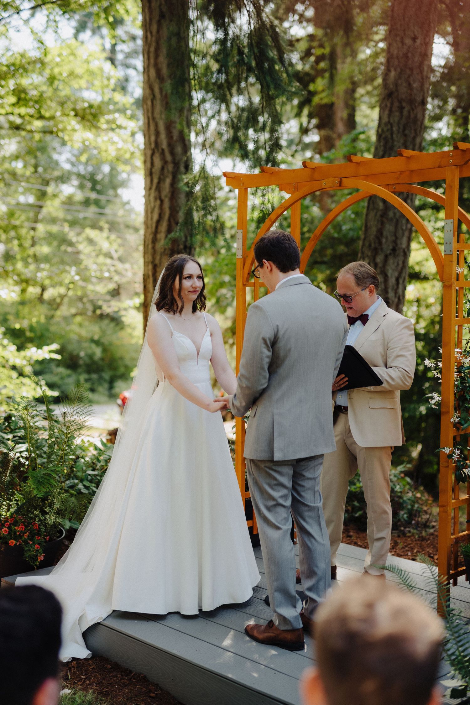 Wedding ceremony takes place under a wooden arch in a lush forest setting with guests seated on white chairs.