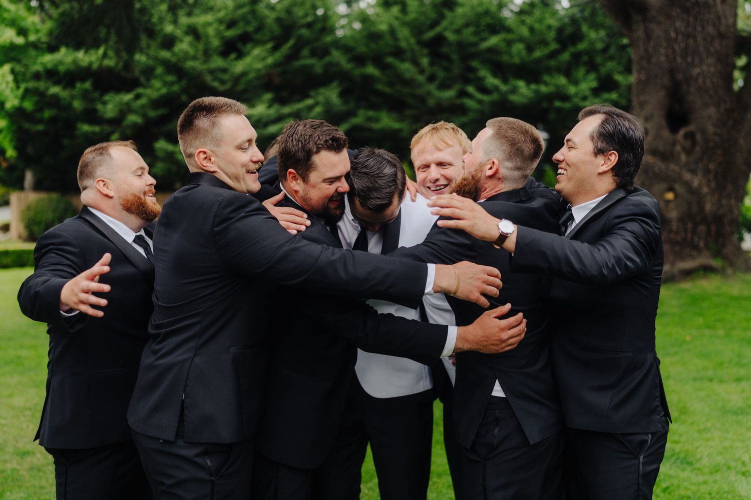 Groomsmen in black suits share a joyful group hug outdoors during wedding celebrations.