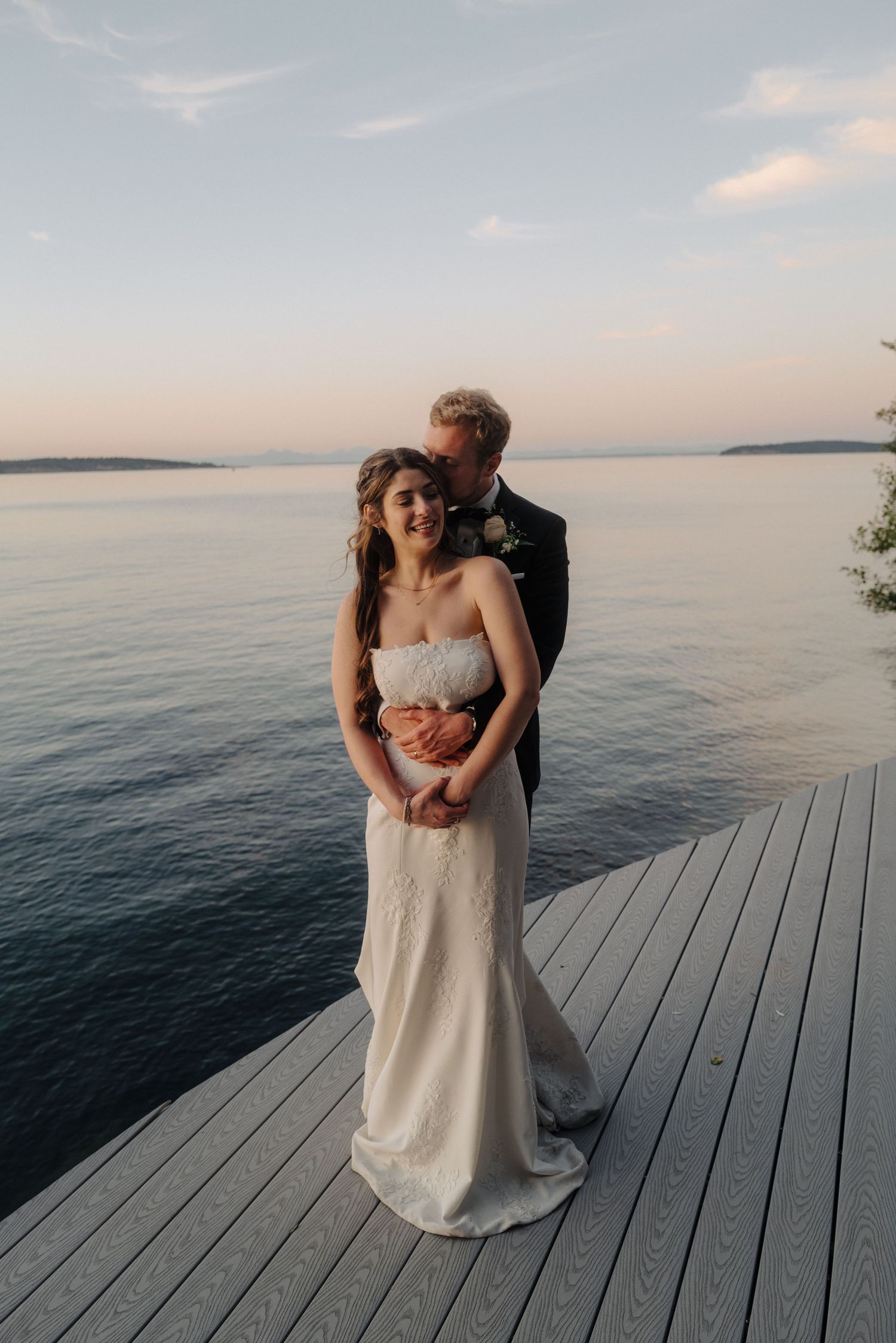 A couple poses together on a wooden dock with calm waters and distant islands at sunset.