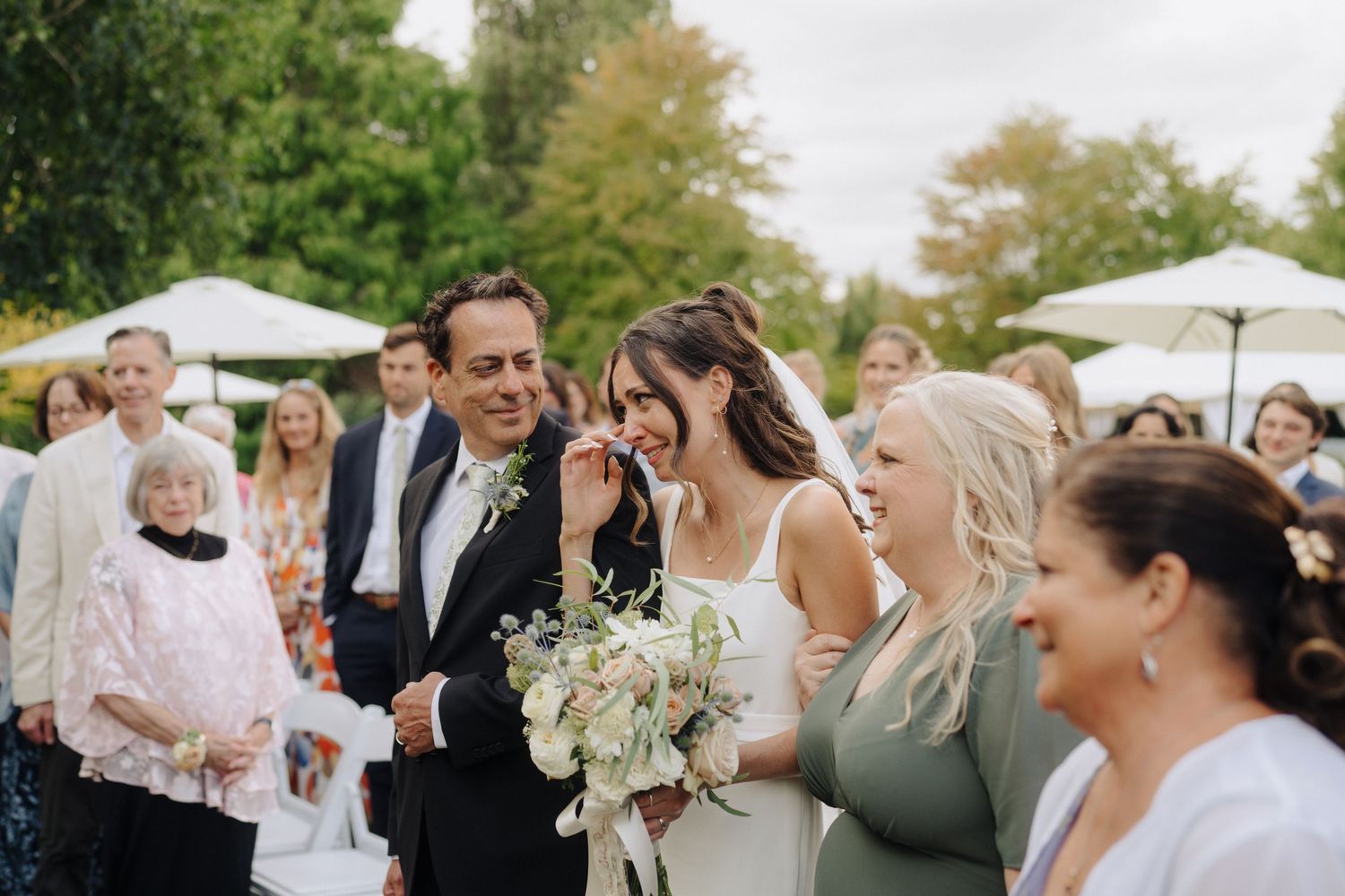 Wedding guests gather outdoors under white umbrellas during a sunny celebration.