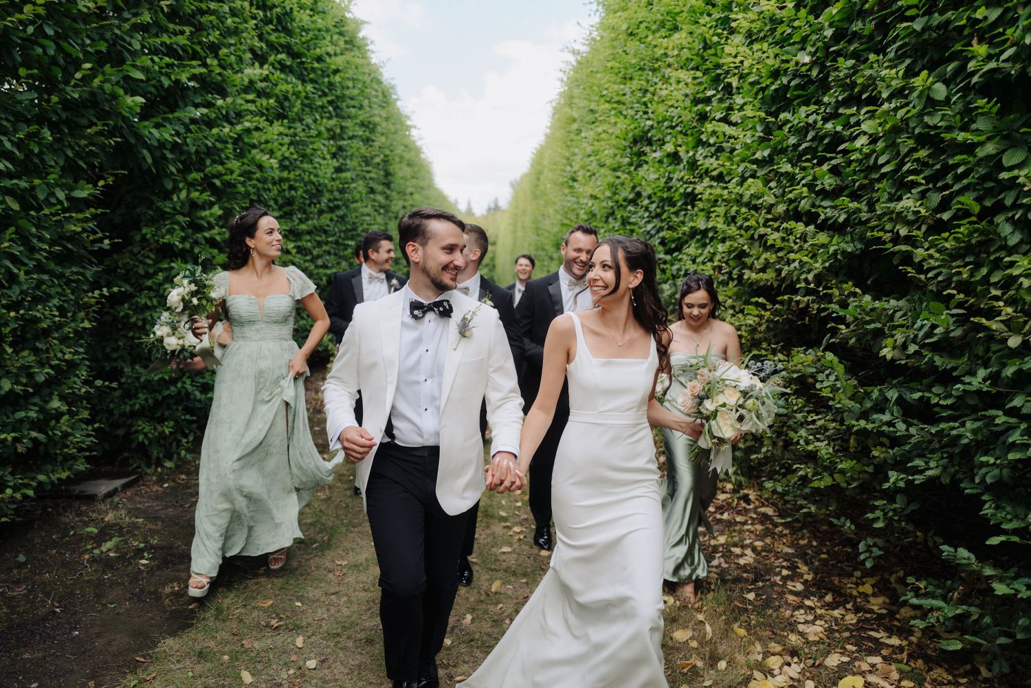 Wedding group strolling through a manicured garden path surrounded by tall hedges.