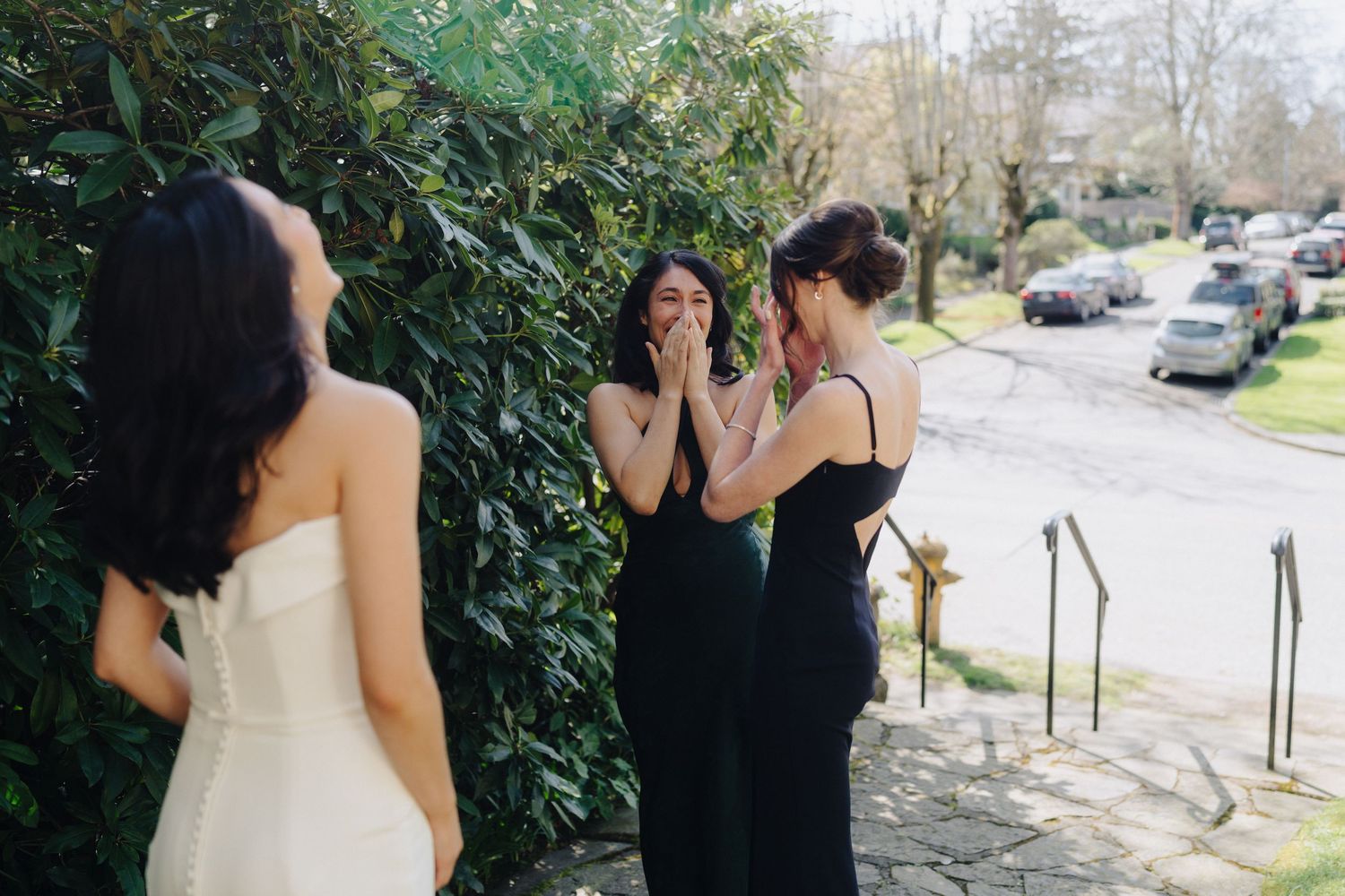 People in formal attire gather and share a lighthearted moment together outside near greenery and parked cars.