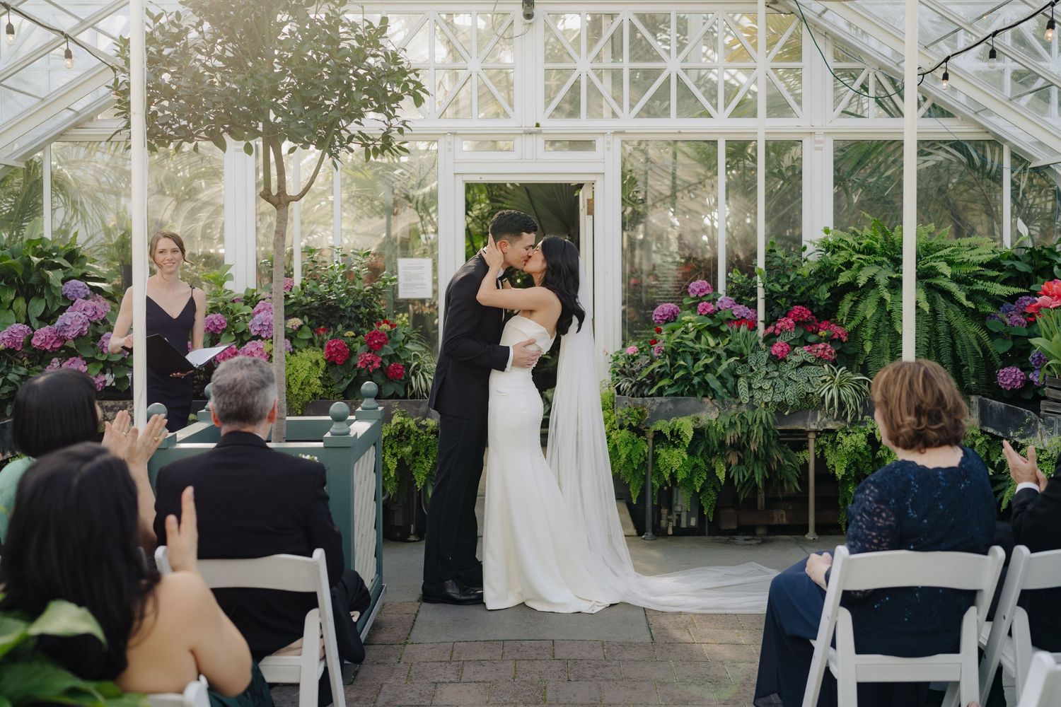 A greenhouse wedding ceremony with guests seated as the couple exchanges vows among vibrant floral displays.