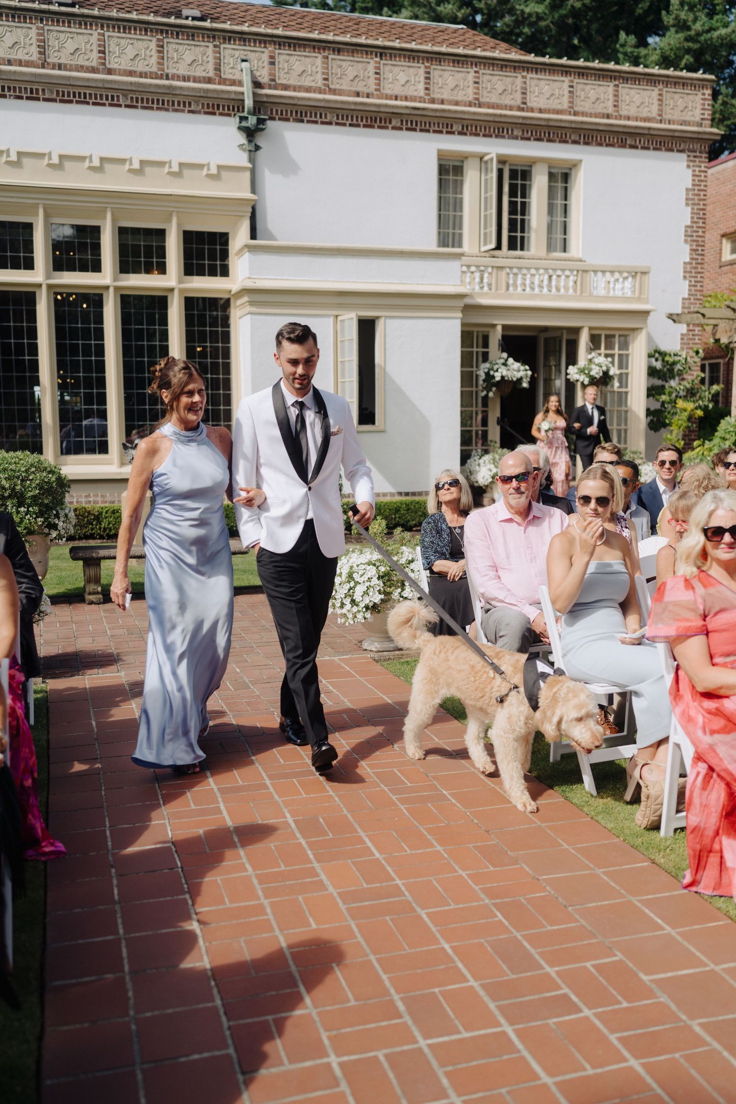 Wedding guests enjoying festivities on a terrace with brick pavers and white building backdrop.