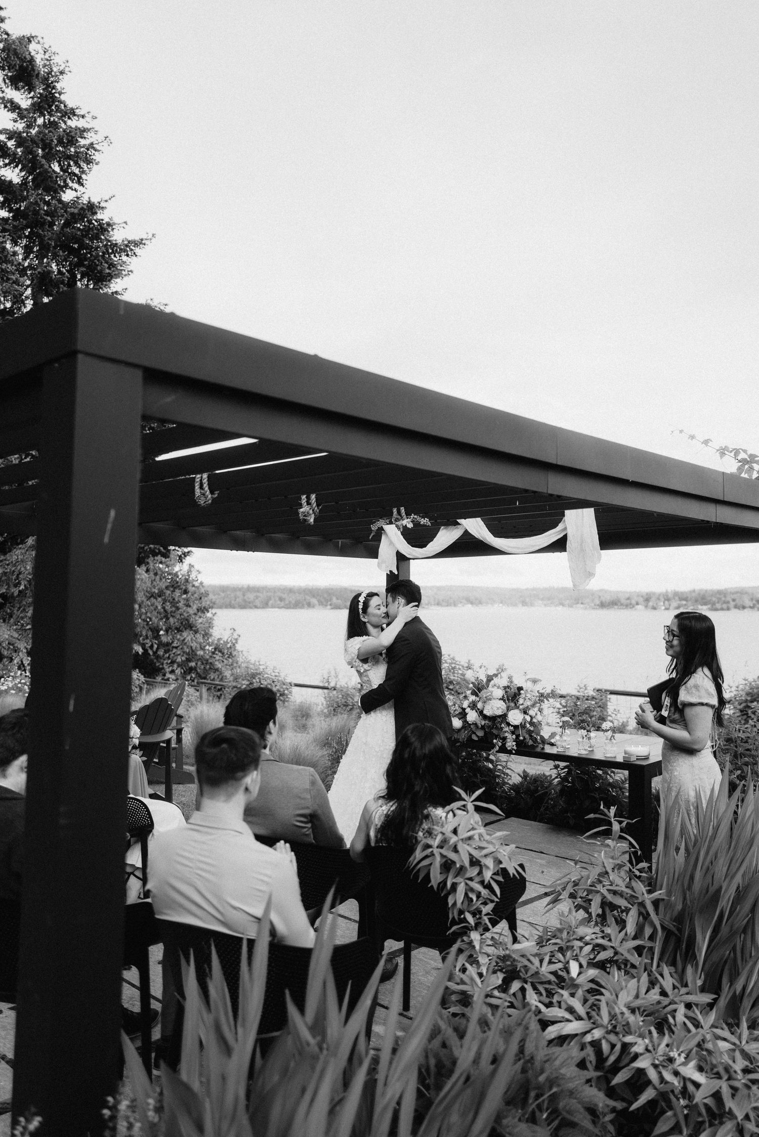 Wedding ceremony guests seated outdoors with water views and decorative plants.