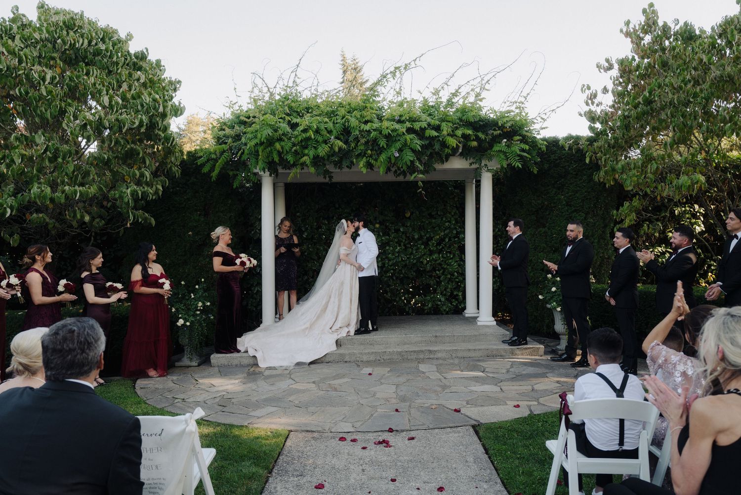 Wedding ceremony in progress at an elegant garden venue with white pillars.