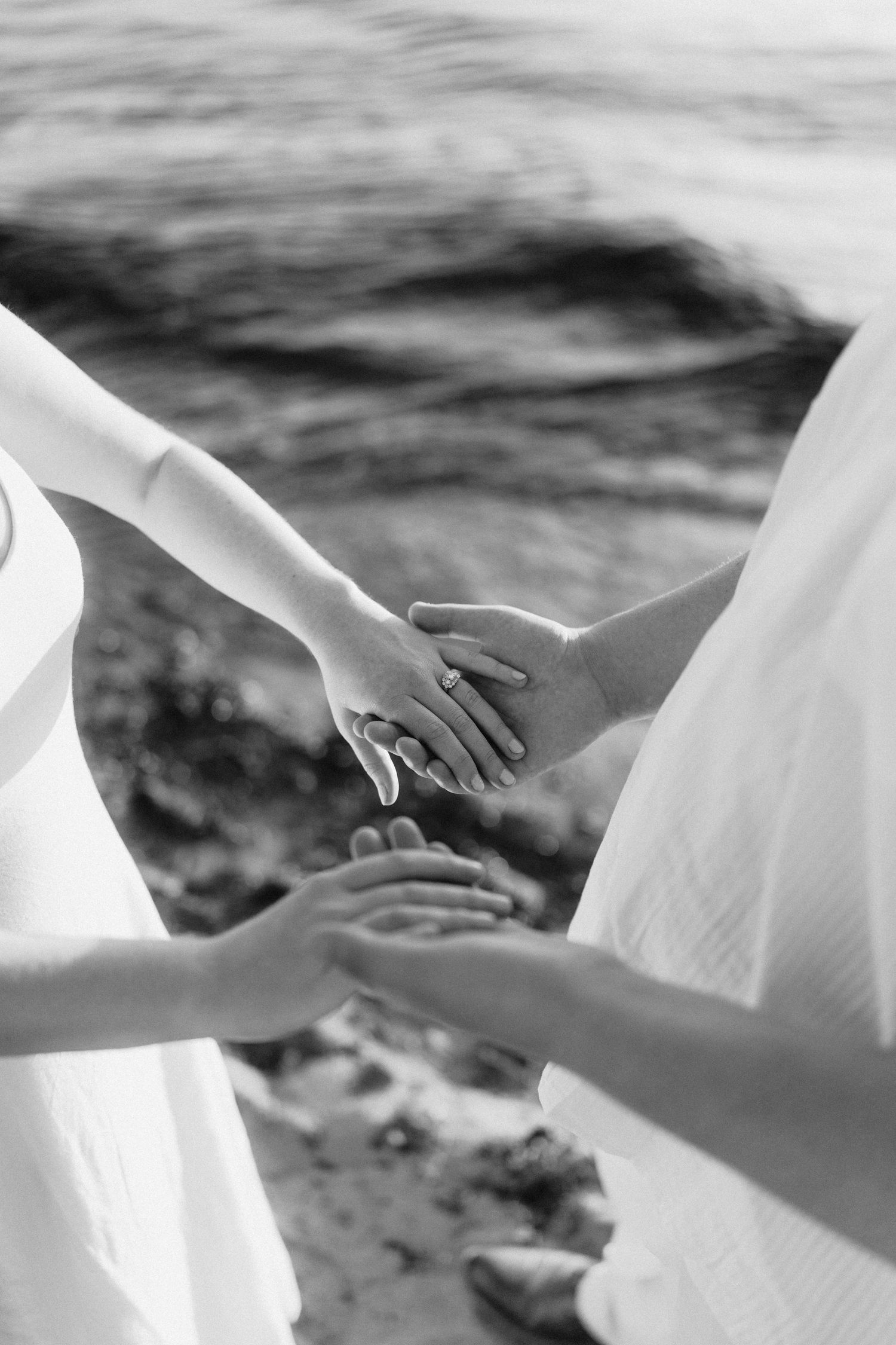 Close-up of hands gently touching against a blurred beach background in black and white.