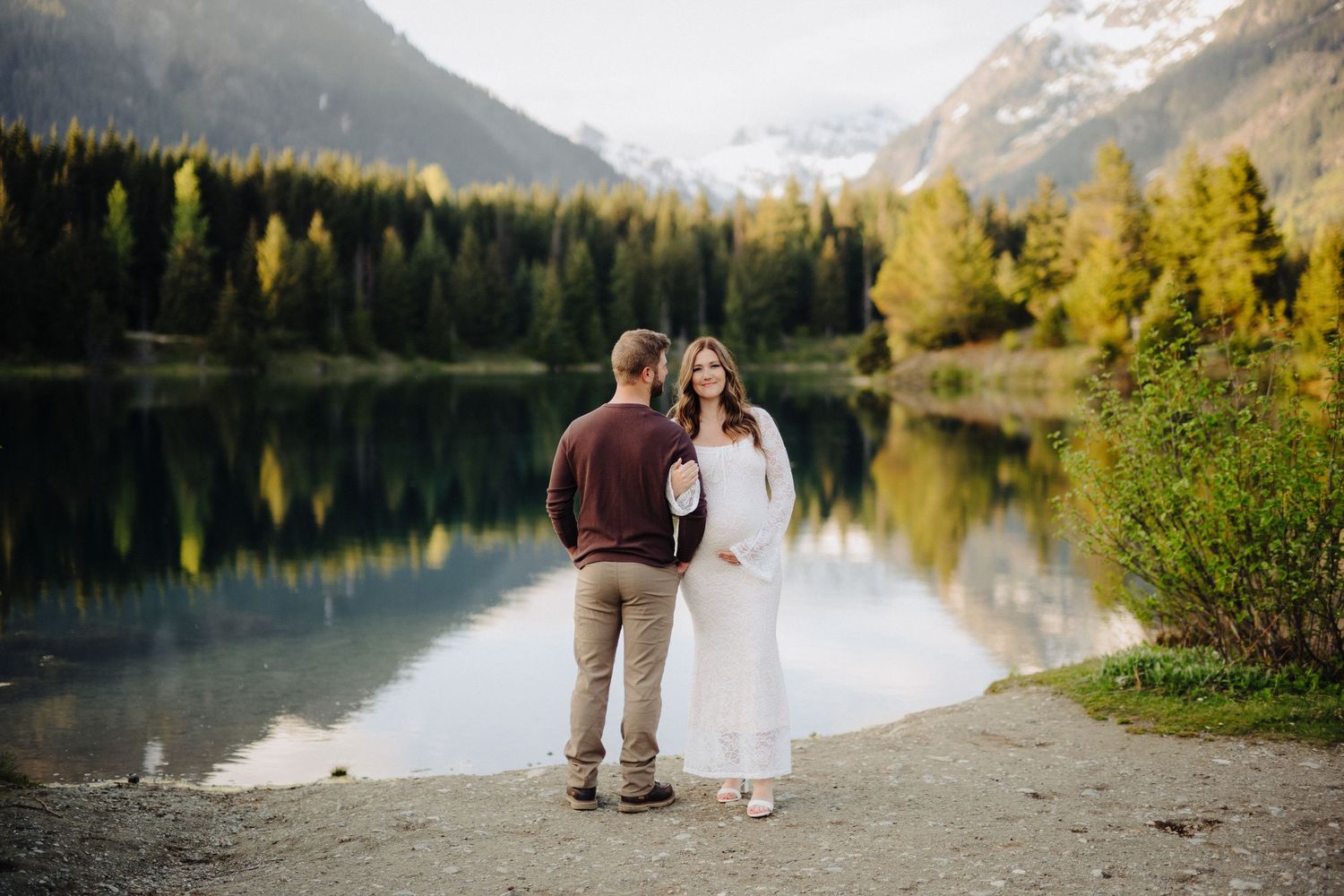 Couple walks along mountain lake shore with snow-capped peaks and evergreen forest in background.