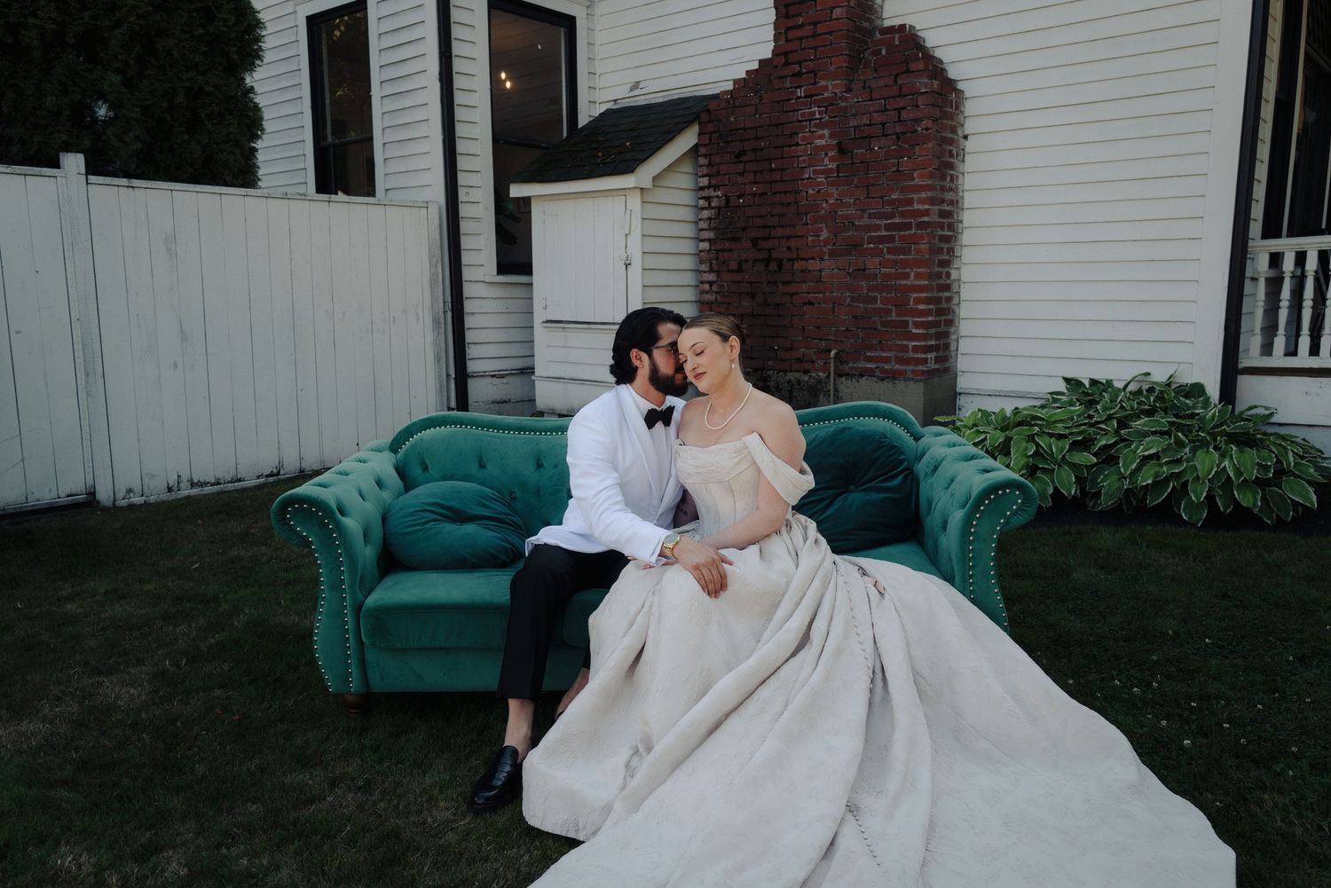 Couple in wedding attire sharing an intimate moment on a teal velvet couch outdoors.