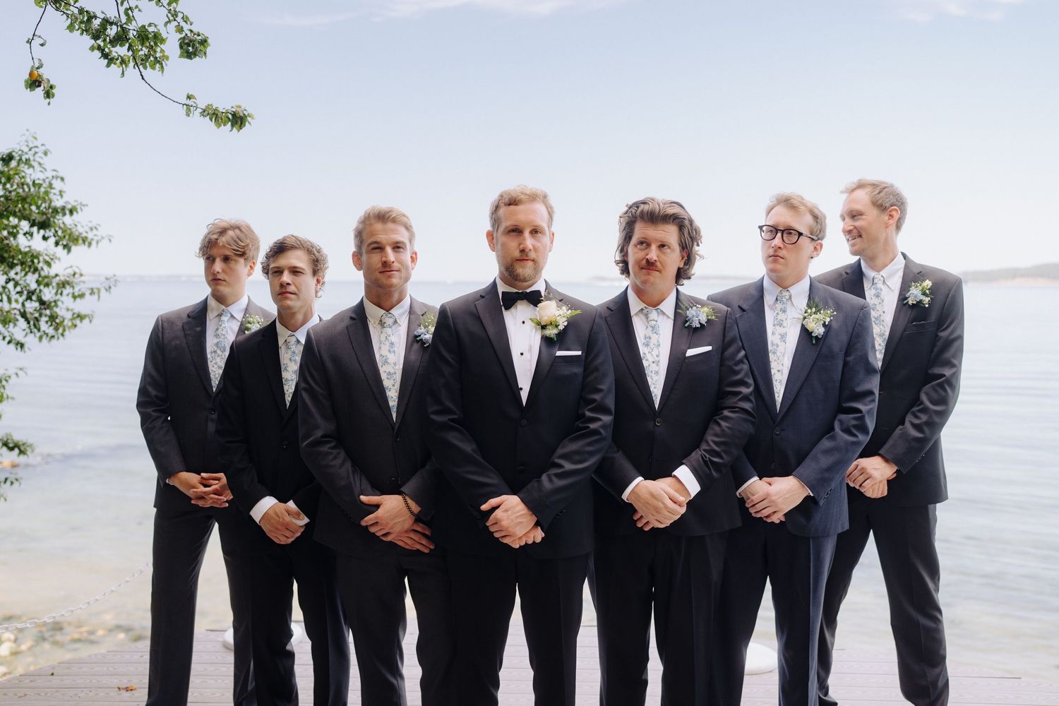 Groomsmen in formal black suits and bowties pose together by the water.
