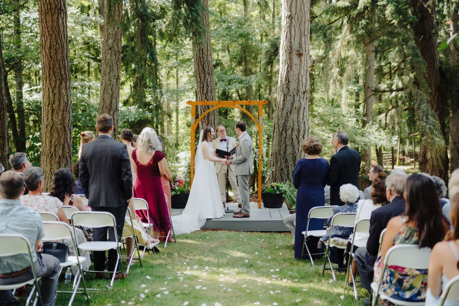 Outdoor wedding ceremony with rows of white chairs set up in a wooded forest area under tall trees.