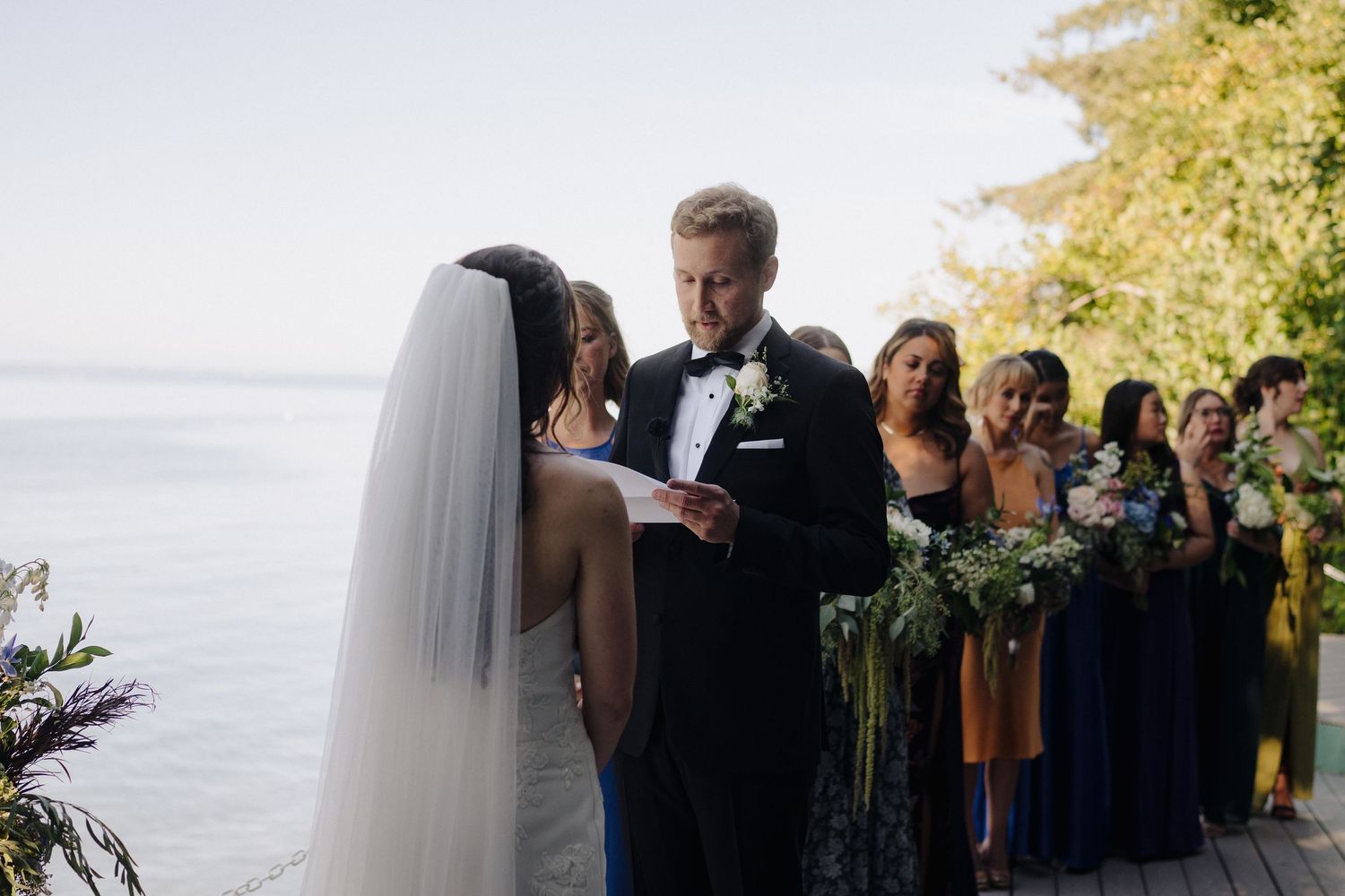 Outdoor wedding ceremony taking place near the ocean with guests watching under golden sunlight.