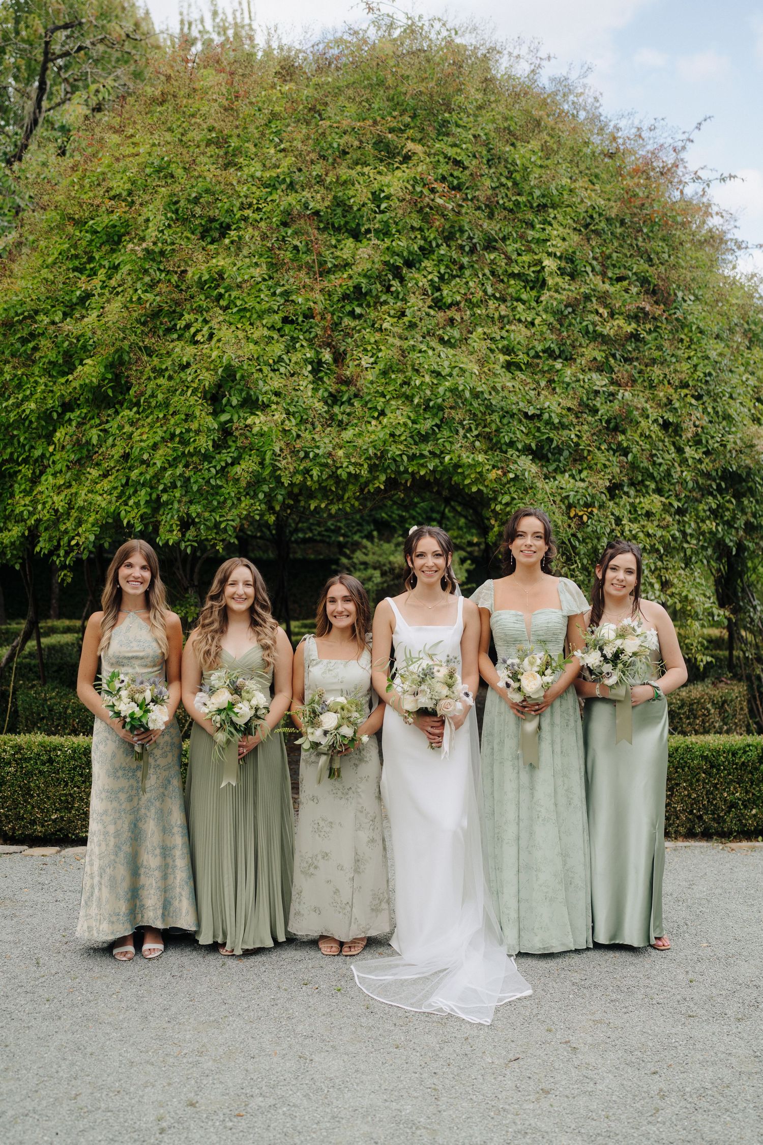 Bridesmaids in sage green dresses pose together with bouquets on a gravel path surrounded by greenery.