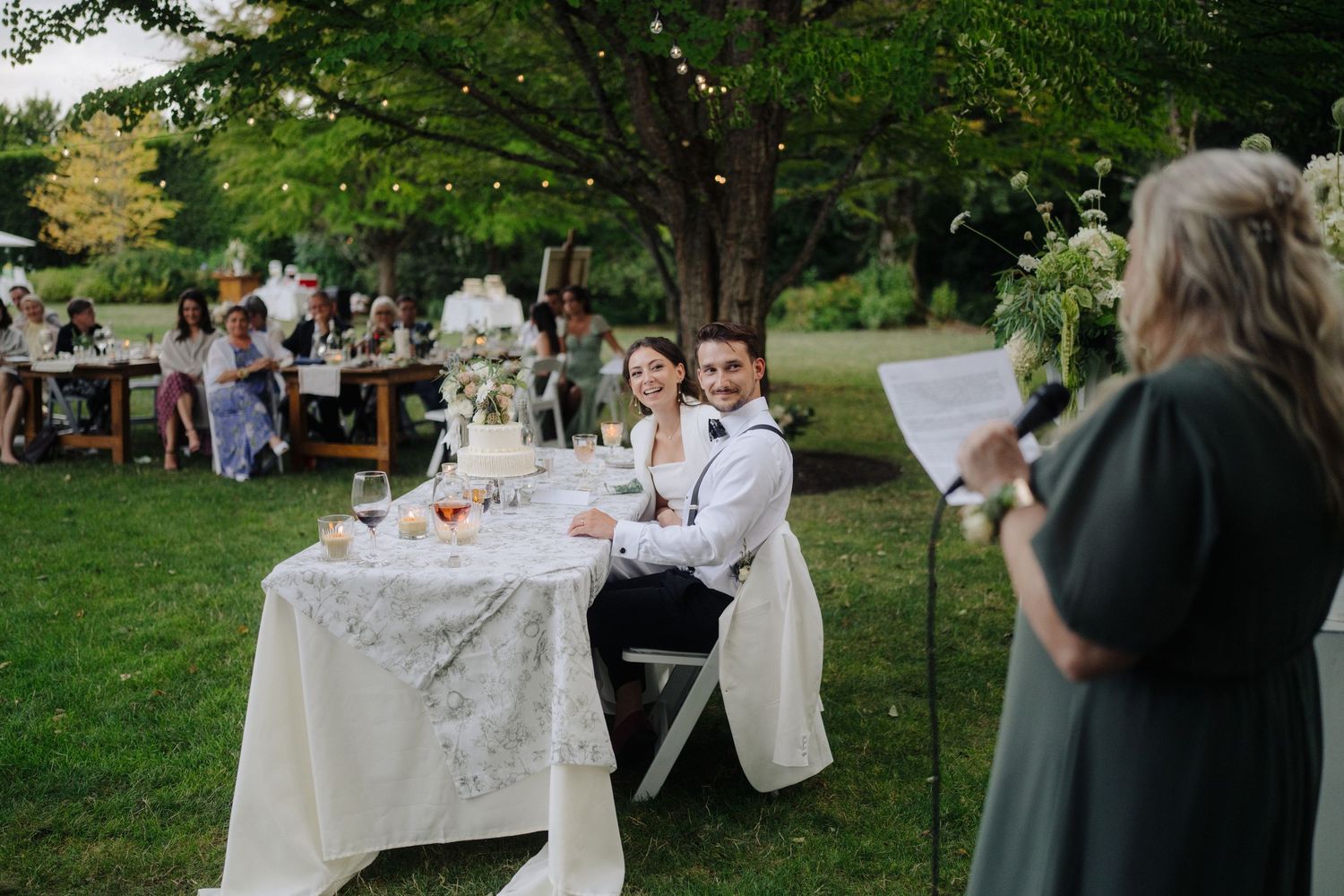 Outdoor wedding reception setup with white tables and decorations under green trees.