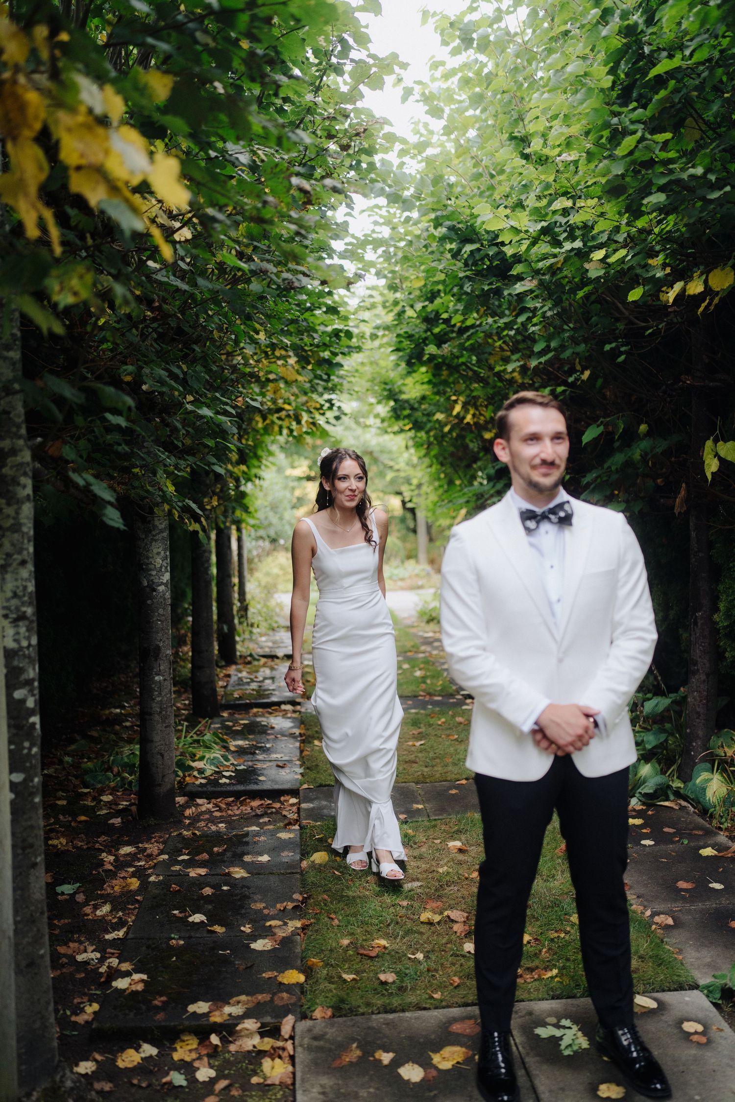 Wedding party poses along a tree-lined path with fallen autumn leaves on the ground.