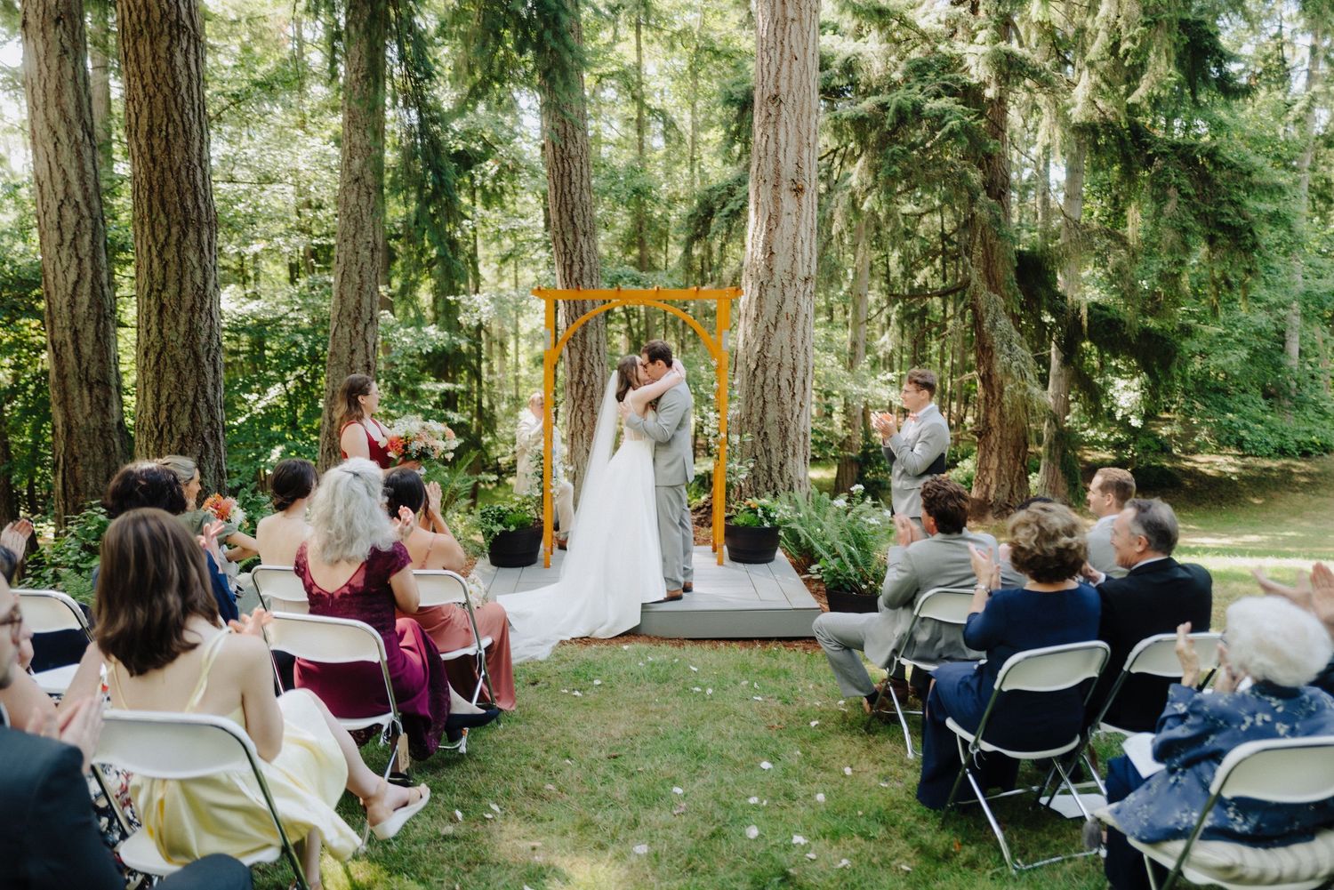Wedding guests seated in white chairs observe an outdoor ceremony among tall evergreen trees.