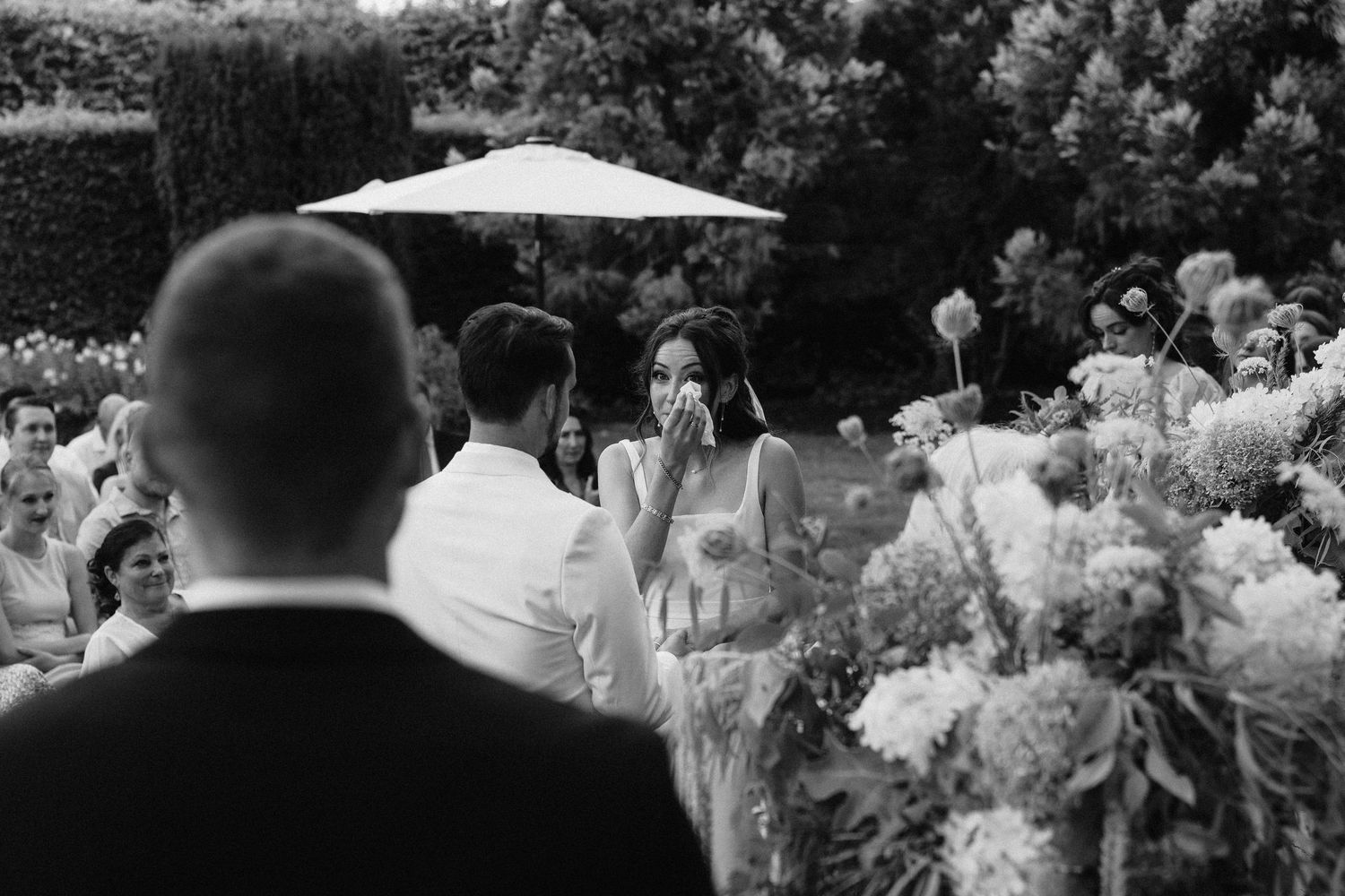 Wedding guests seated in white chairs on a lawn under umbrellas during an outdoor ceremony.