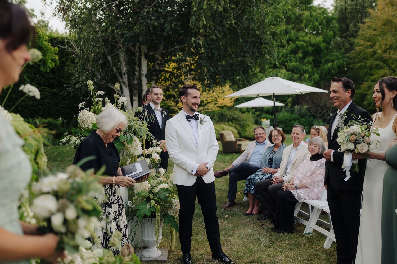 An intimate outdoor garden wedding ceremony with guests seated on white chairs among greenery.