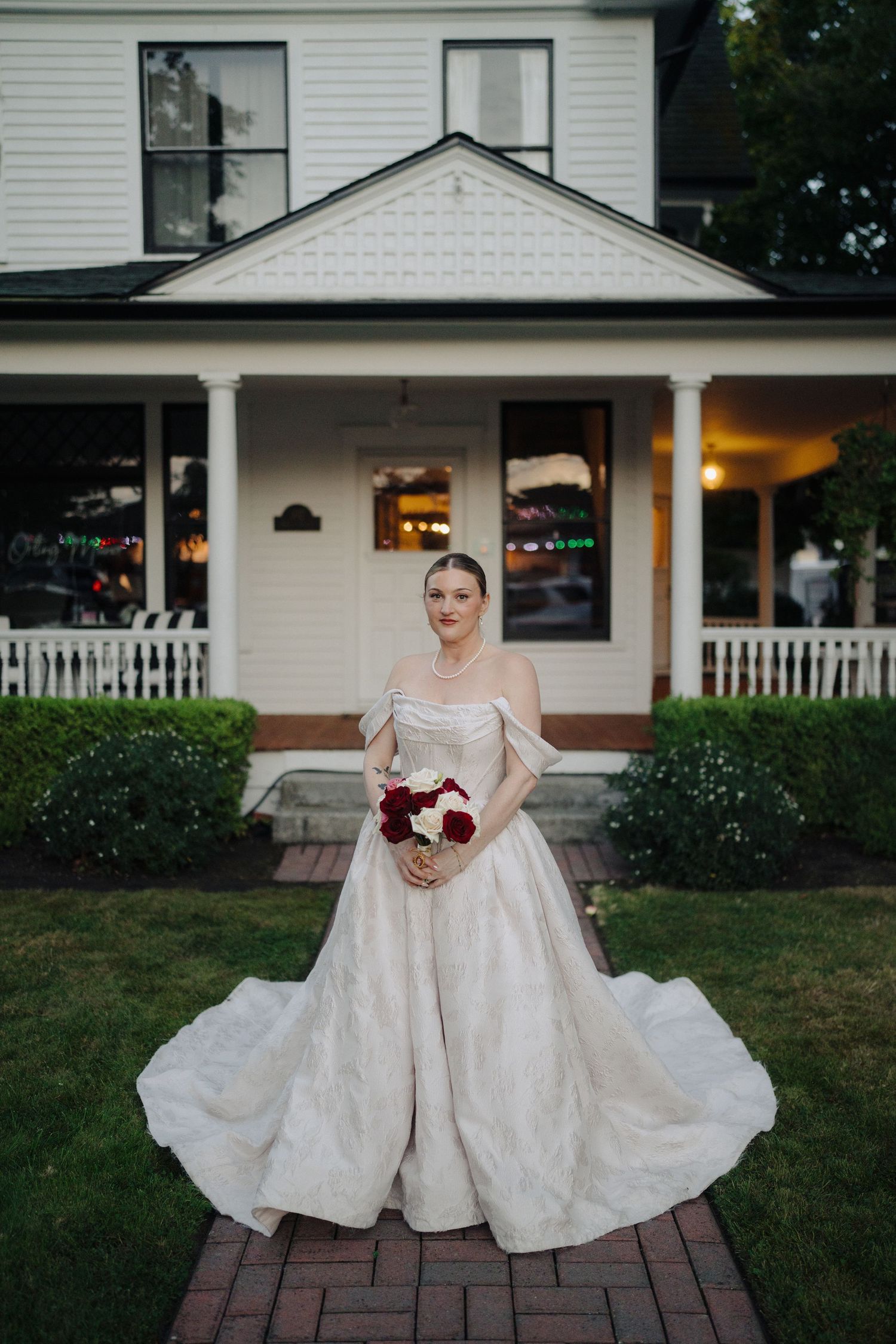 Elegant bride in a classic white ballgown poses on brick pathway in front of white house.