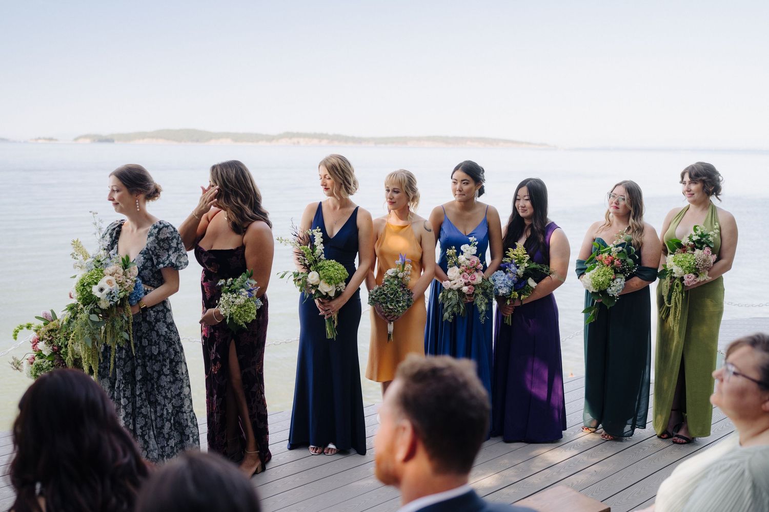 A row of bridesmaids in blue and green dresses stand together outdoors at a wedding celebration.
