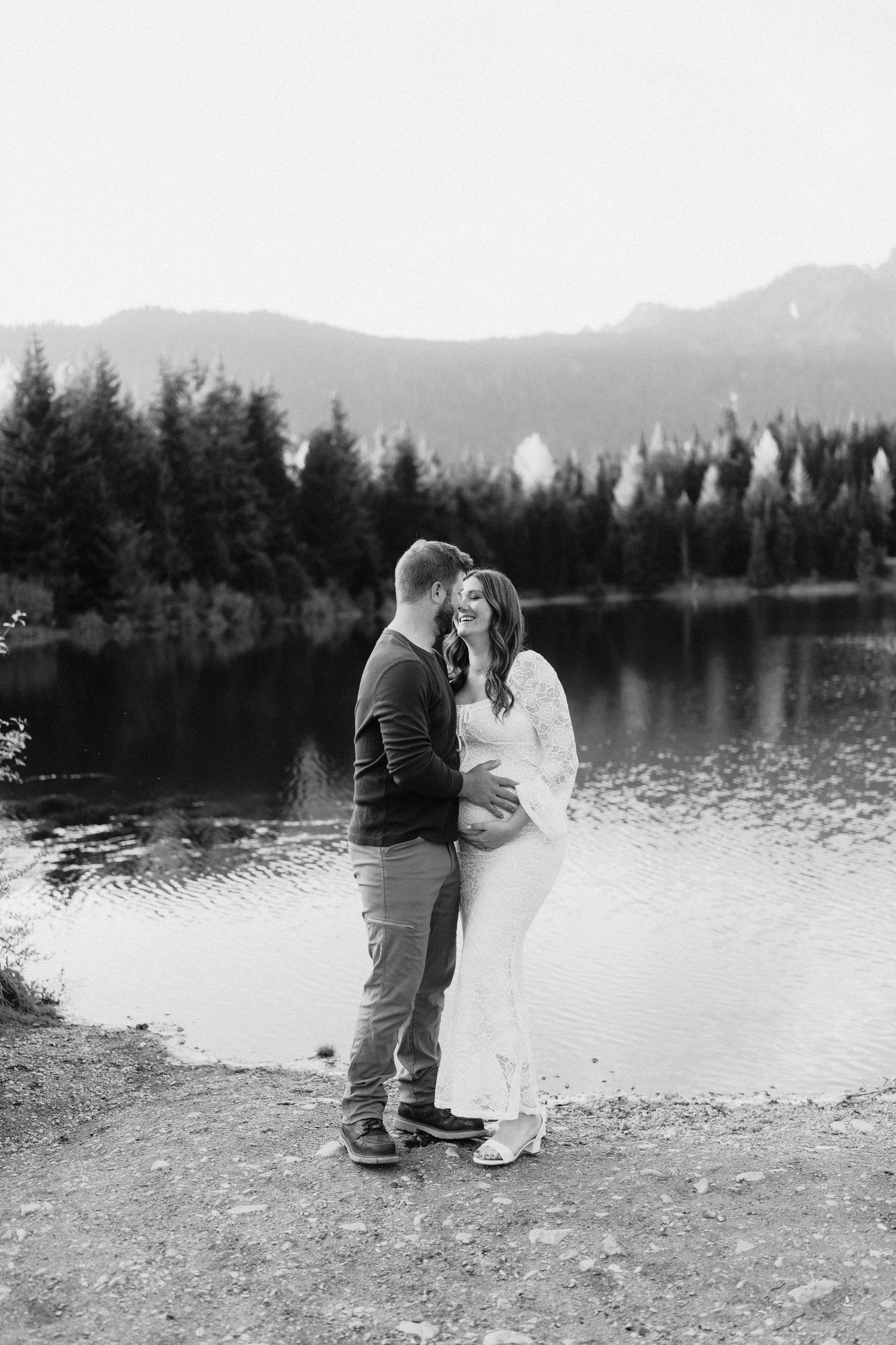 Black and white photo of a couple embracing on a lakeside beach with mountains and forest in the distance.