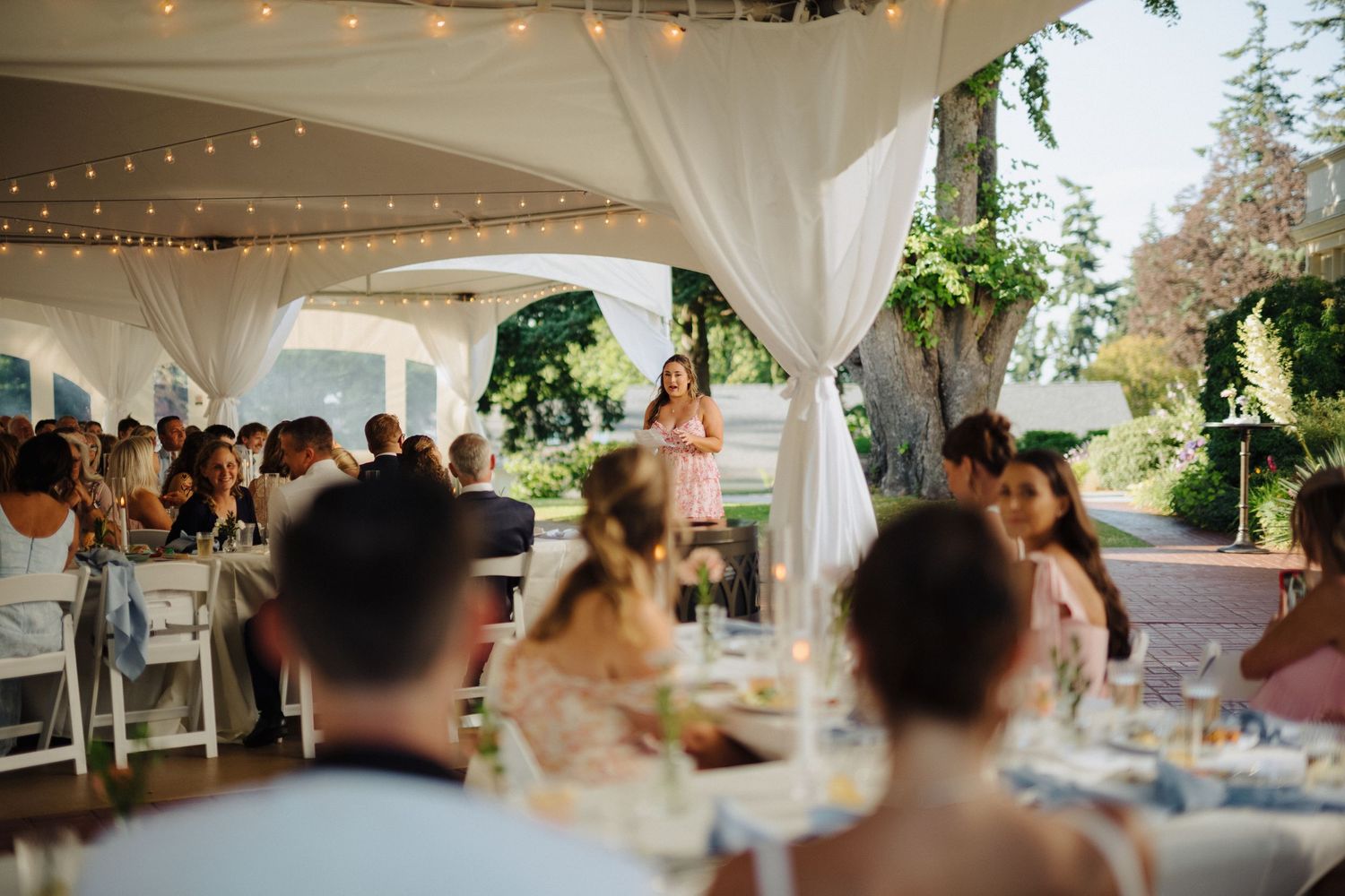 Wedding reception under a white tent with string lights and guests seated at decorated tables.