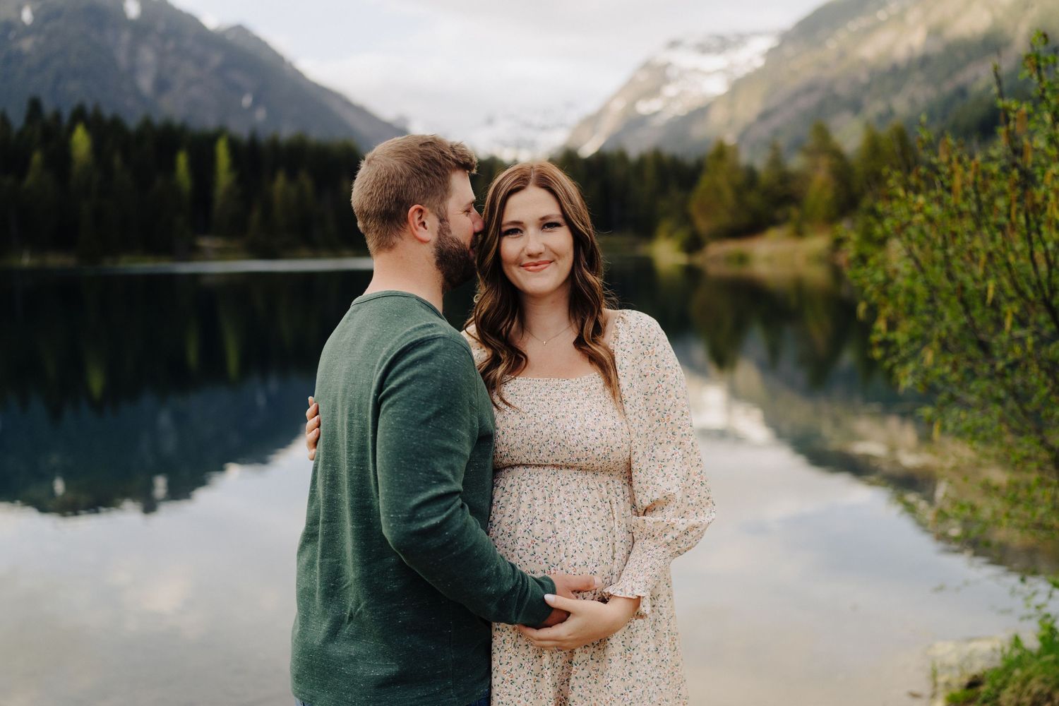 Couple embraces by a serene mountain lake surrounded by evergreen forests and snow-capped peaks.