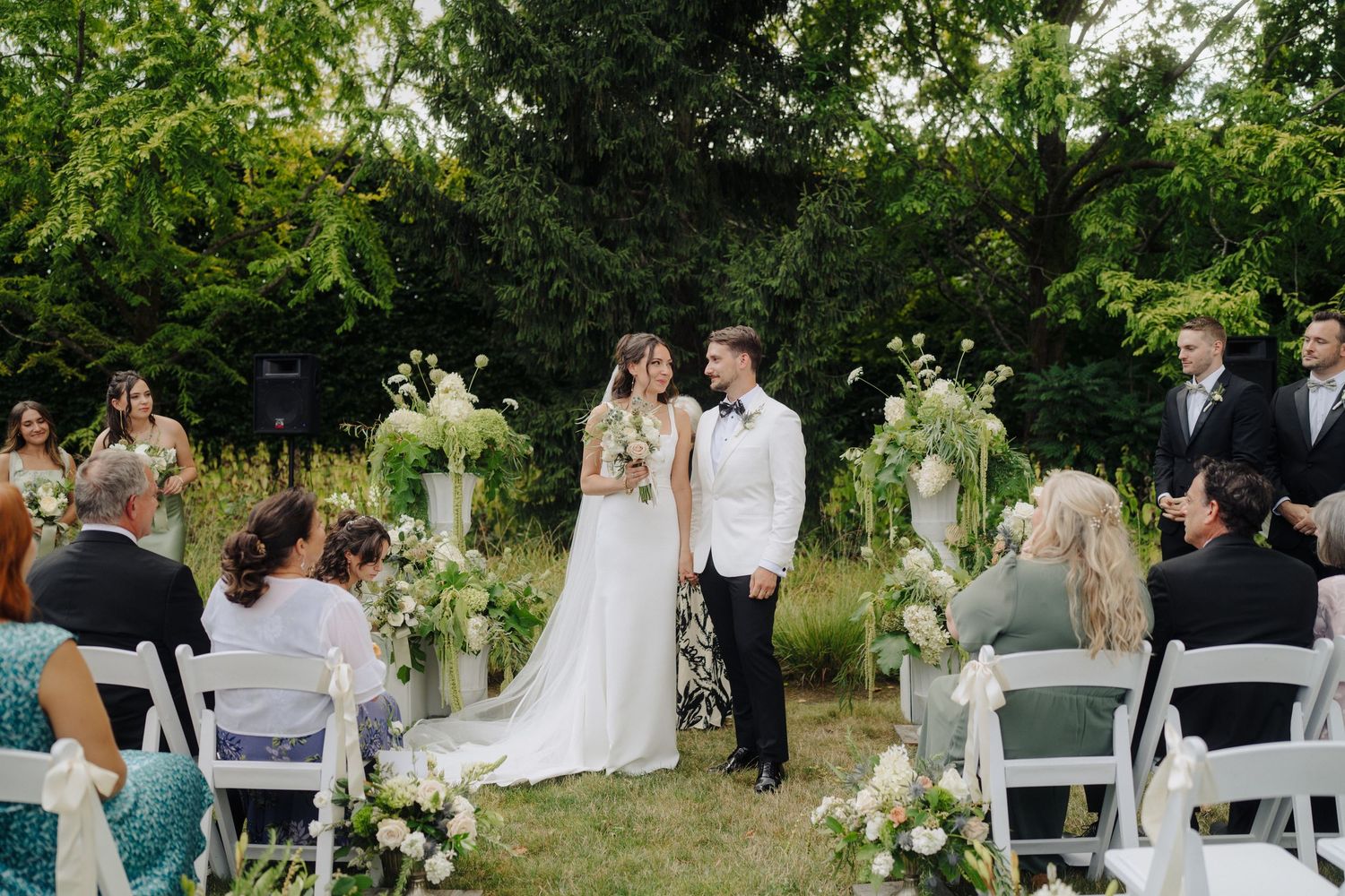 Outdoor wedding celebration with white chairs arranged in rows among lush garden scenery.
